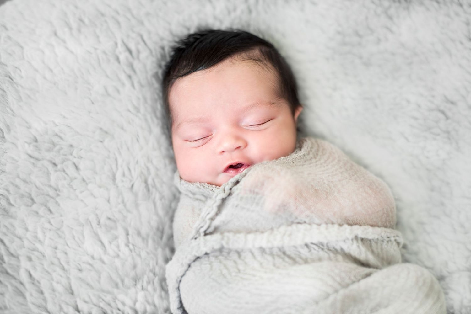 A sleeping newborn baby swaddled in white fabric lies on a soft white blanket in a studio setting.