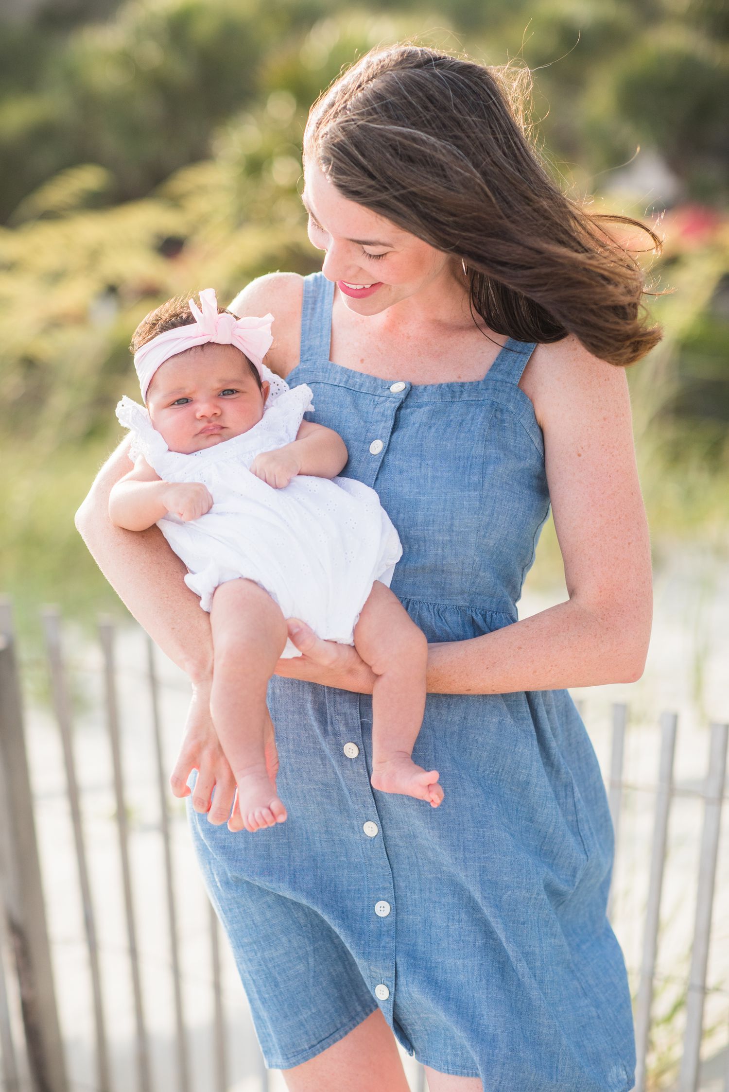 Woman in denim dress holds baby in white outfit against beach fence backdrop.St. Augustine Beach, Hammock Beach photographer.