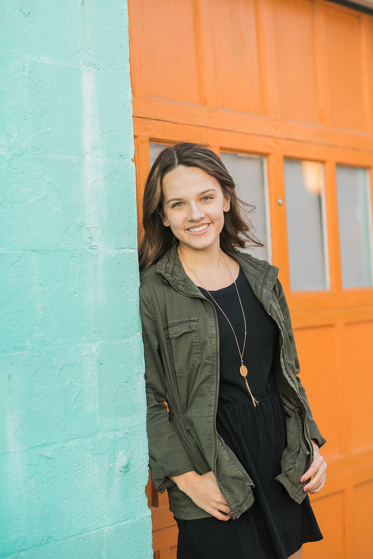 A casual portrait against a vibrant orange and turquoise wall wearing an olive green jacket and black top.