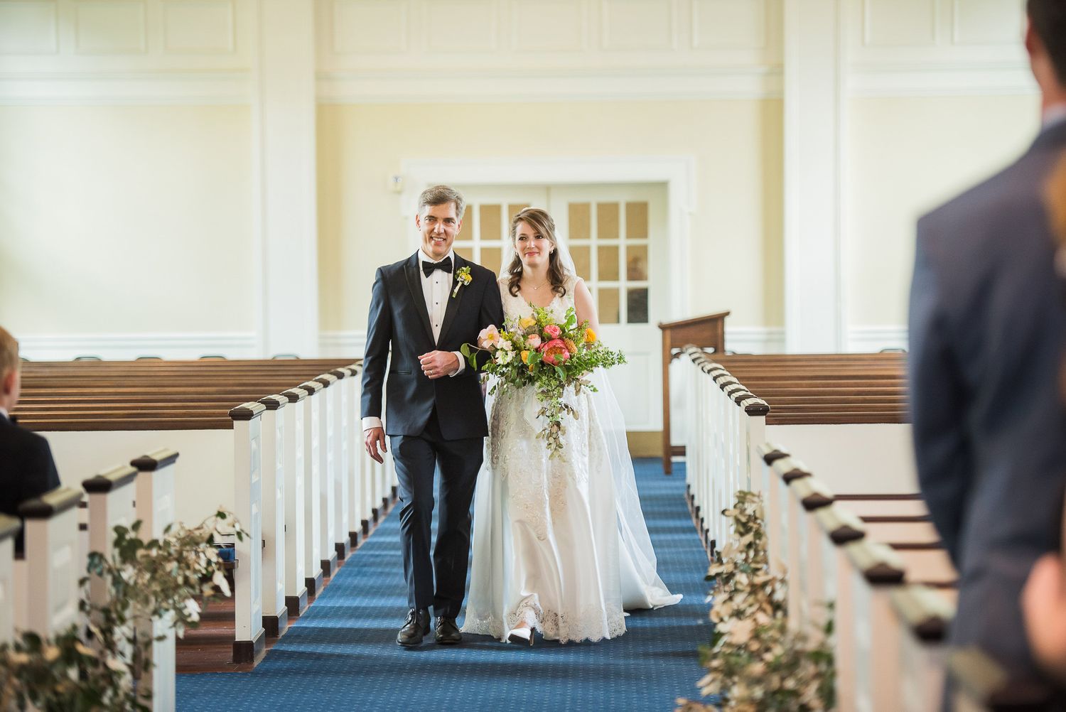 A wedding party enters a bright church sanctuary decorated with floral arrangements along the aisle.