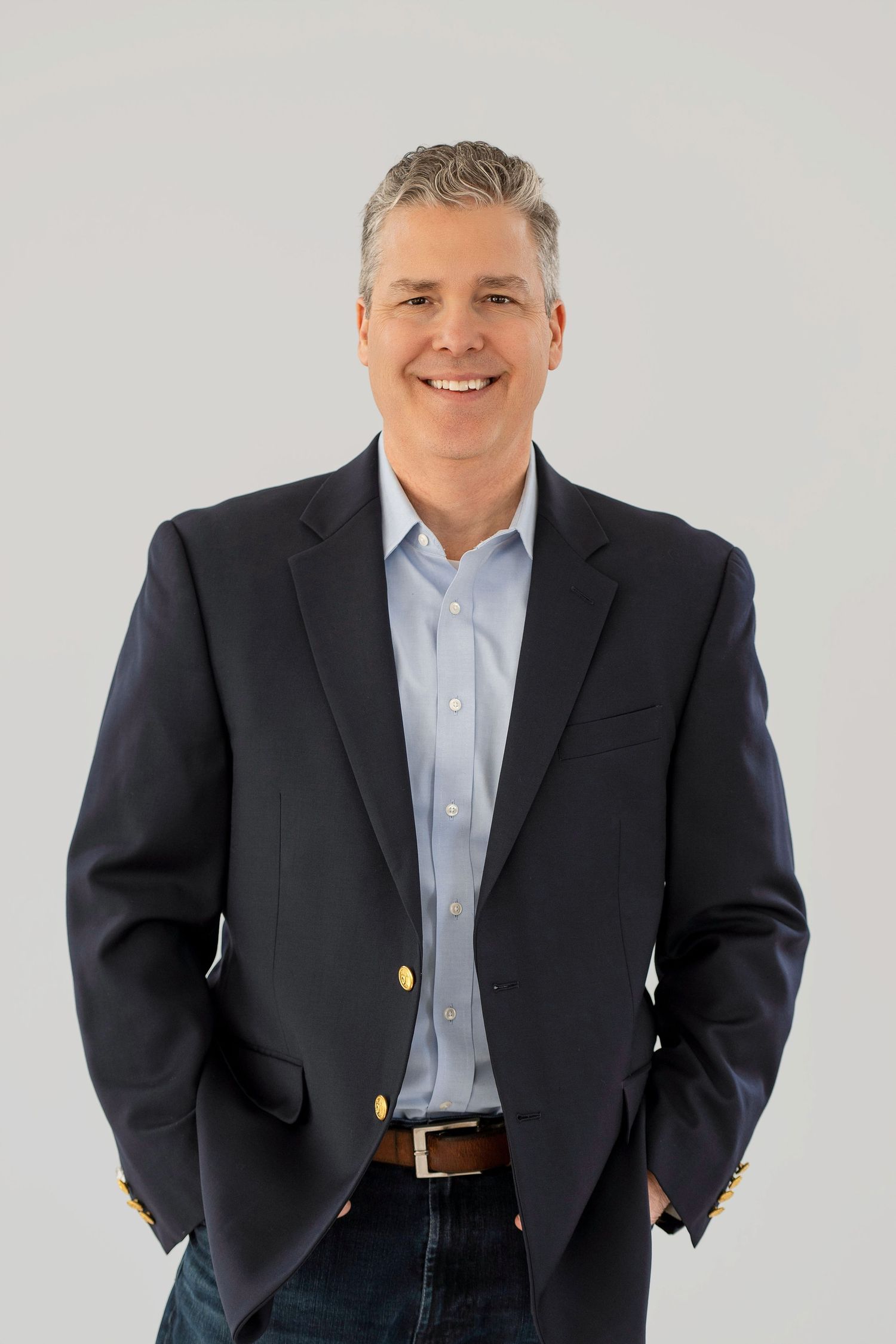 Professional studio headshot of someone in a dark suit jacket and light blue dress shirt against a white background.