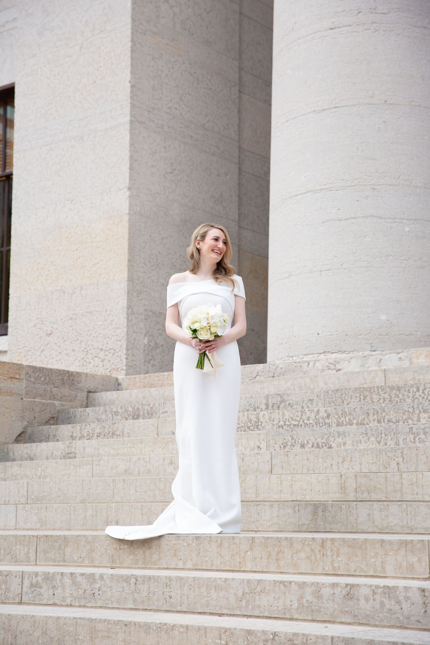 A bride in a white wedding dress sits gracefully on stone steps with a bouquet.