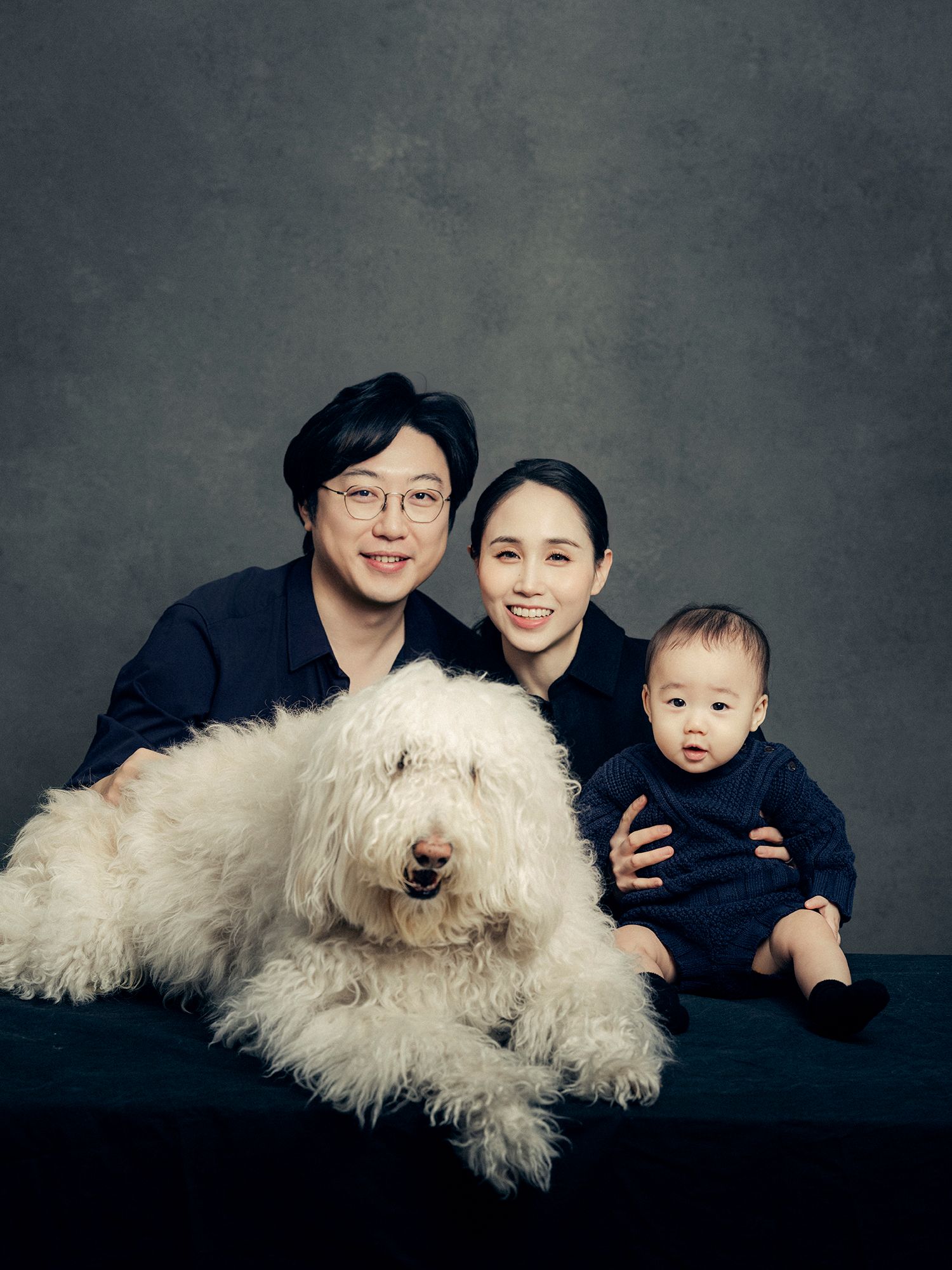 A family portrait featuring a large fluffy white dog posing with three people against a dark gray background.