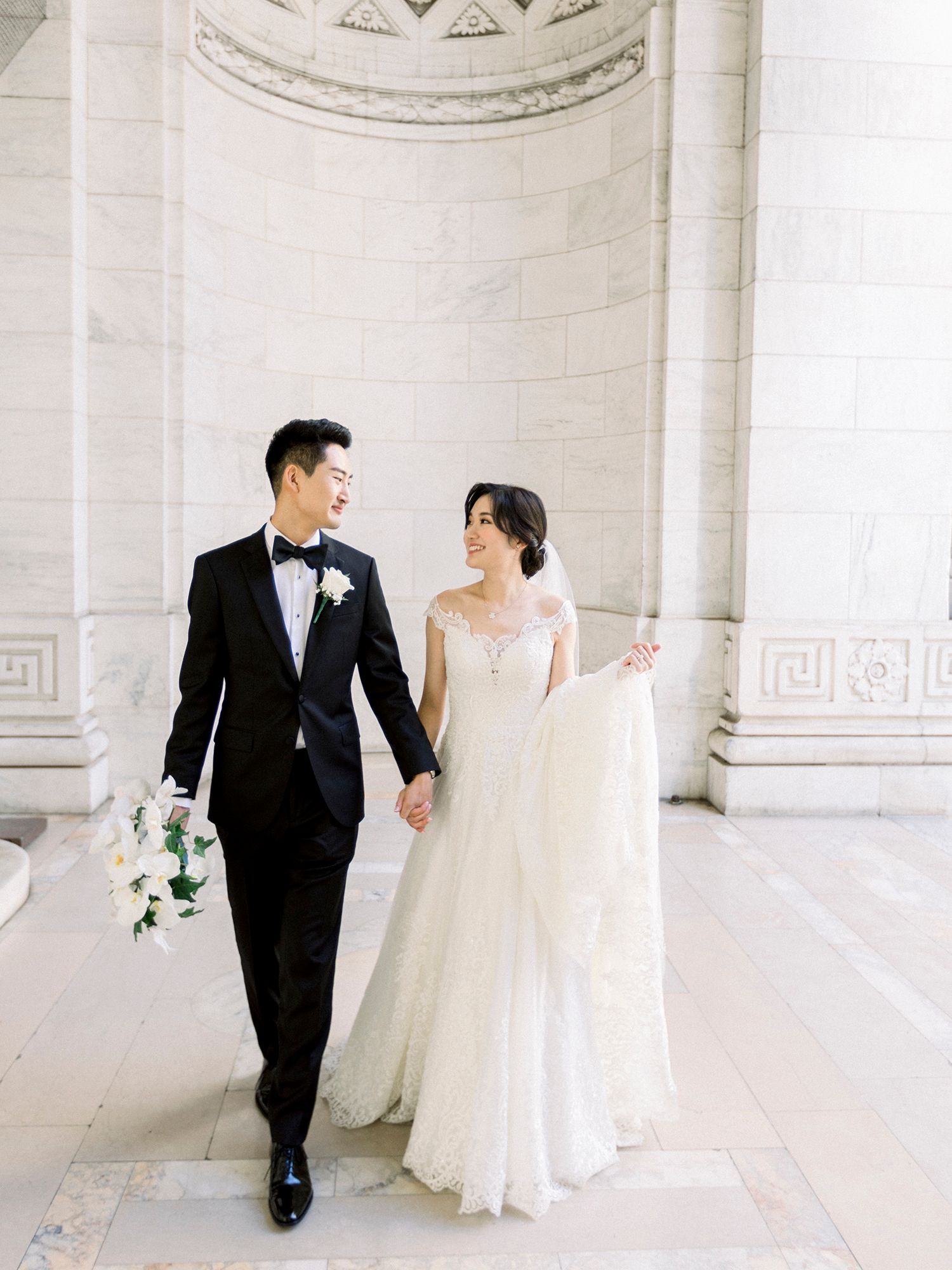 A couple walks hand in hand through an elegant columned hallway in their wedding attire.