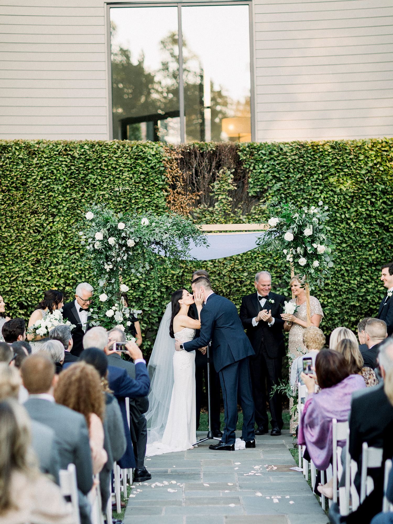 Outdoor wedding ceremony setup with white chairs and floral arrangements against a modern building.