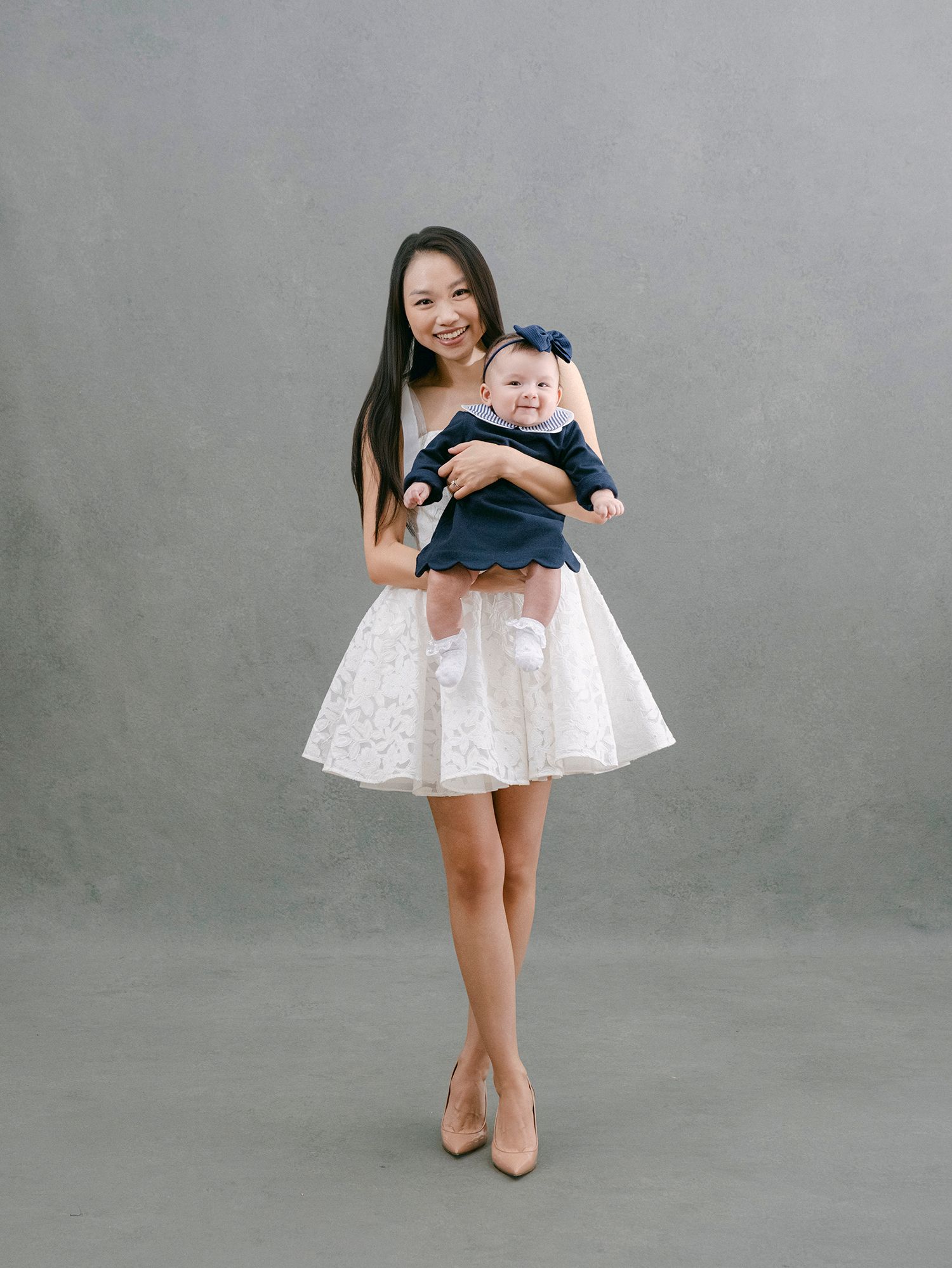 A woman in a white lace dress holds a baby wearing navy blue against a gray studio backdrop.
