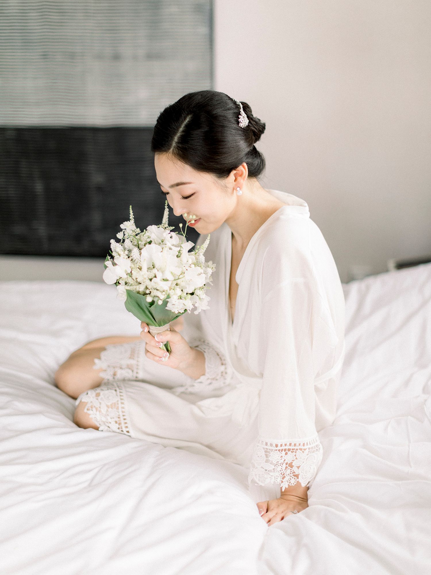 Woman in white robe sits elegantly on bed with white flowers and soft morning light.