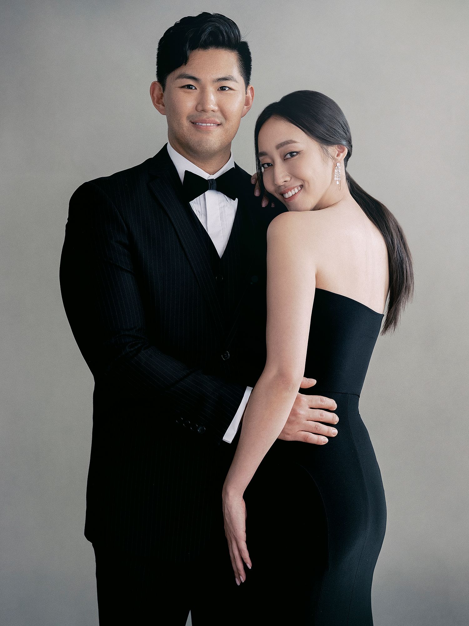 A couple in formal black attire poses together against a neutral grey studio background.