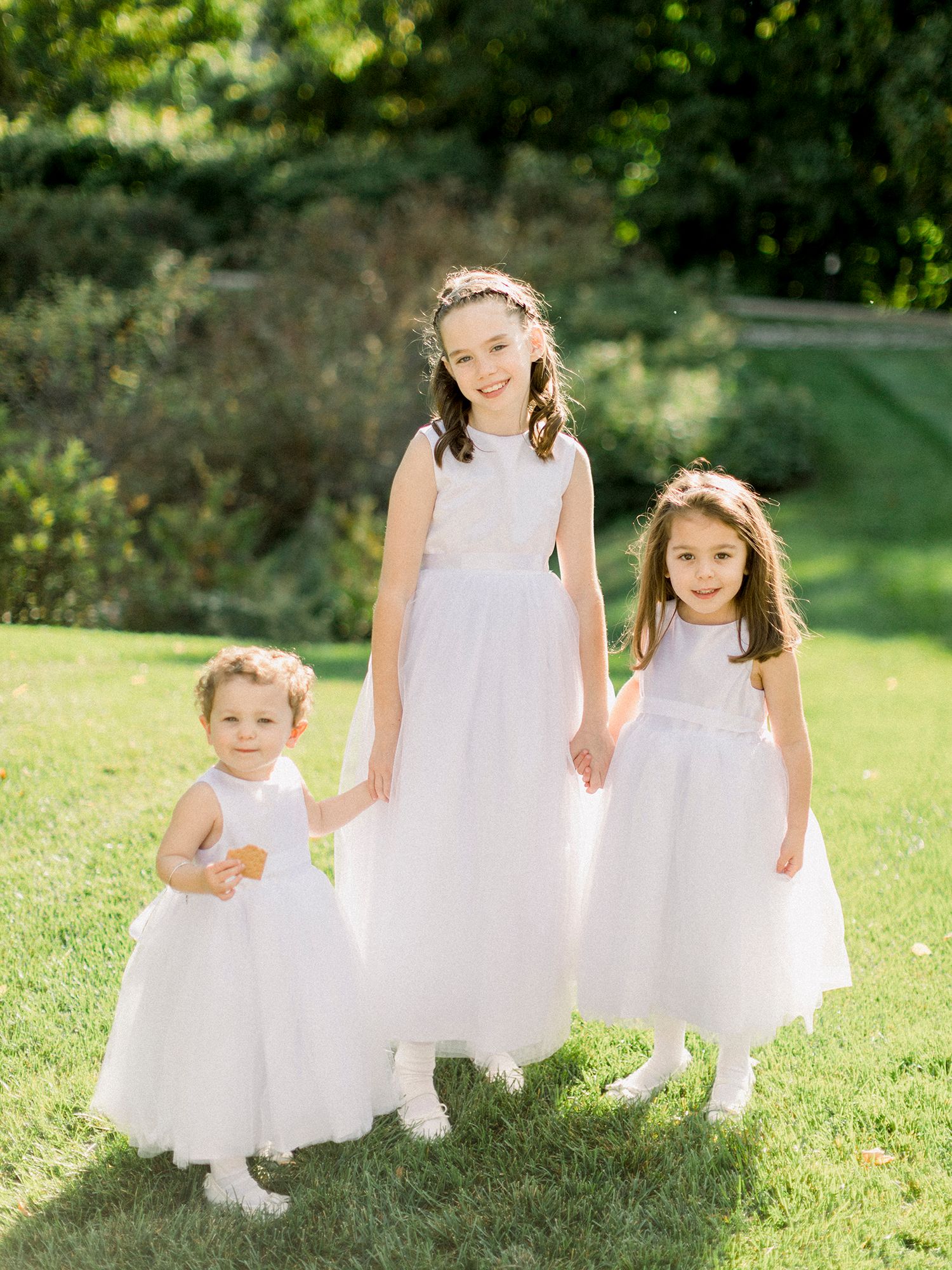 Adorable flower girls in white dresses pose together on sunny garden lawn.