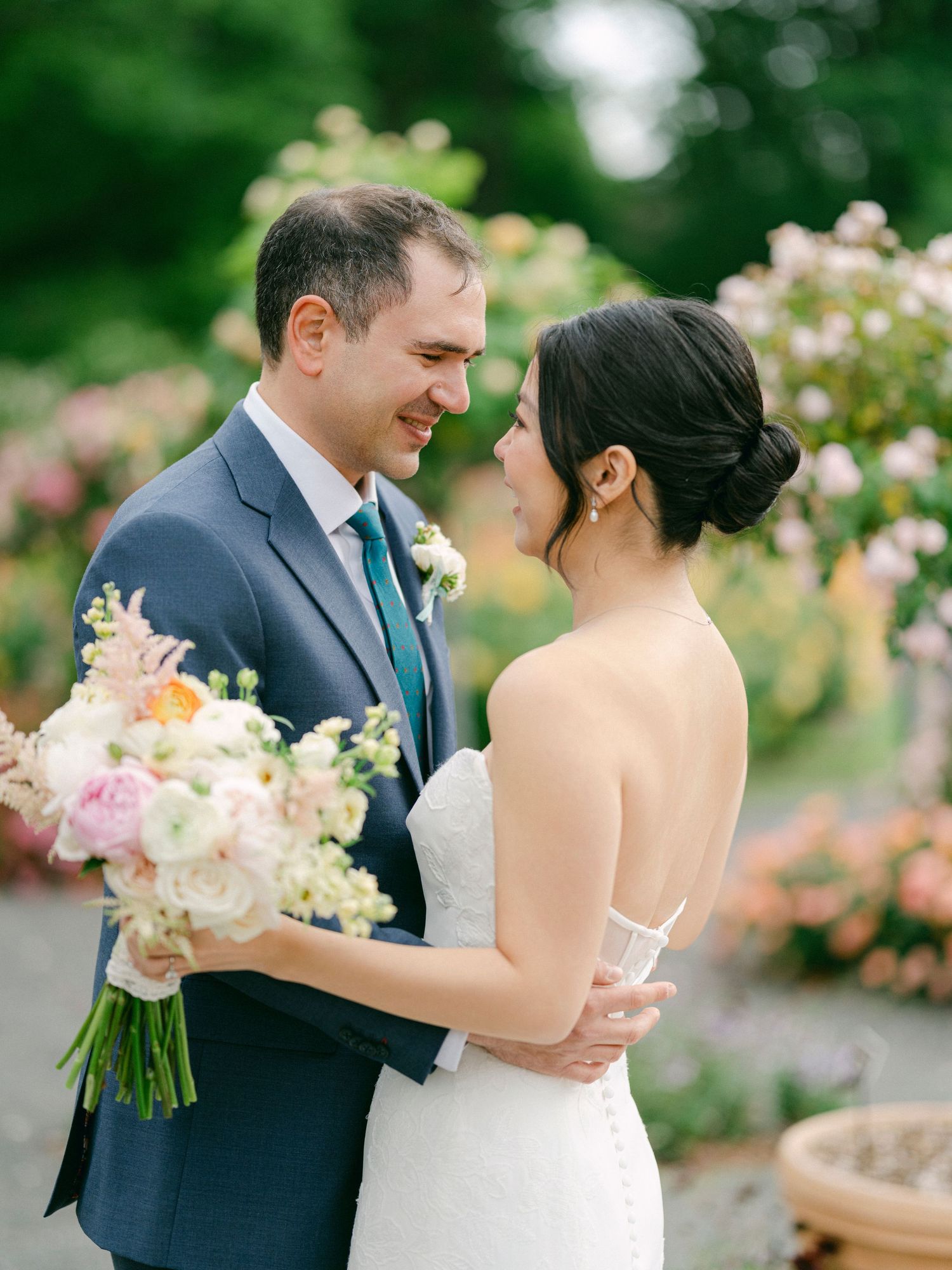 Joyful wedding couple shares intimate moment in rose garden with bridal bouquet.