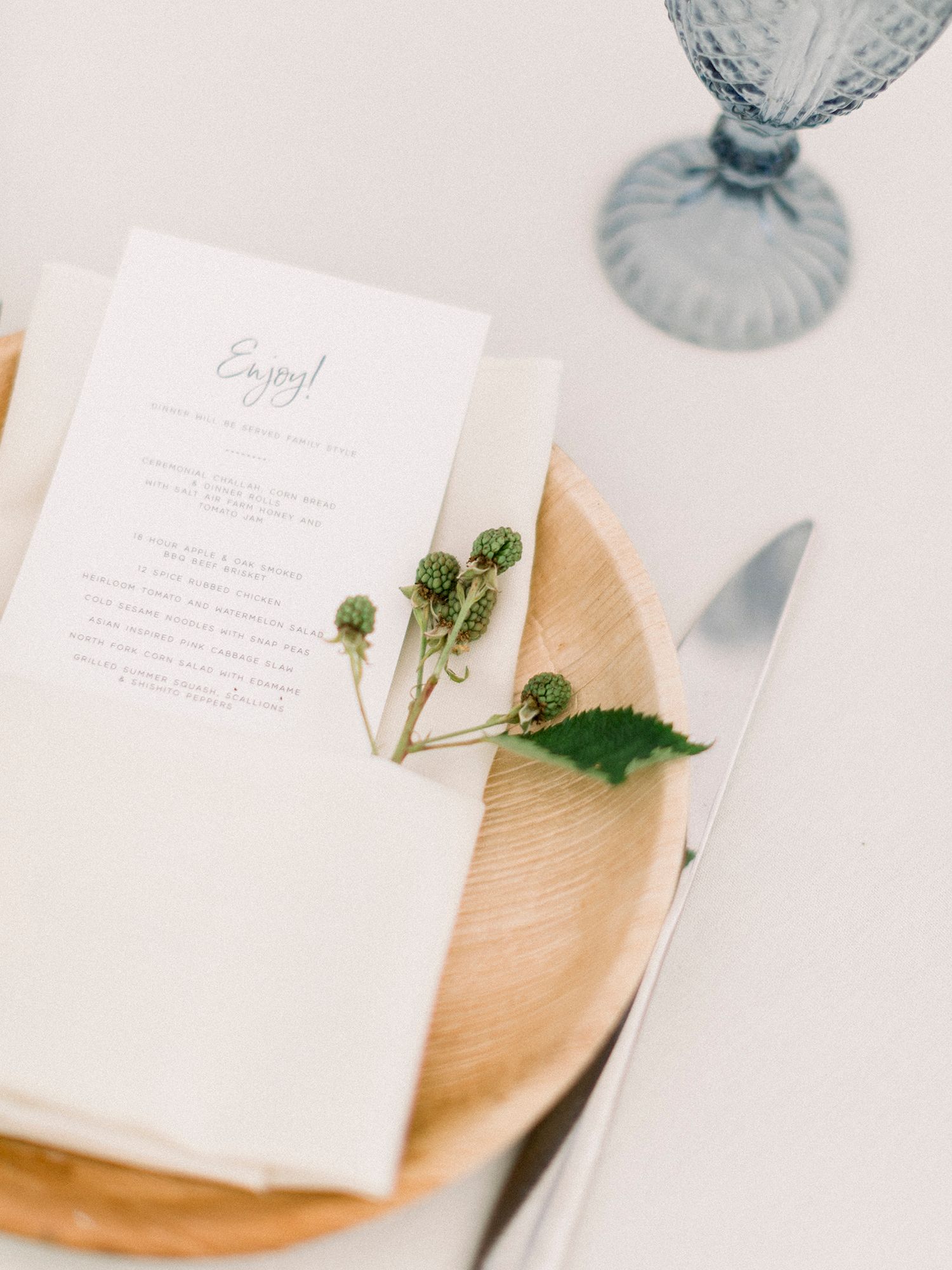 Elegant wedding menu on wooden plate with blue glassware and botanical accents.