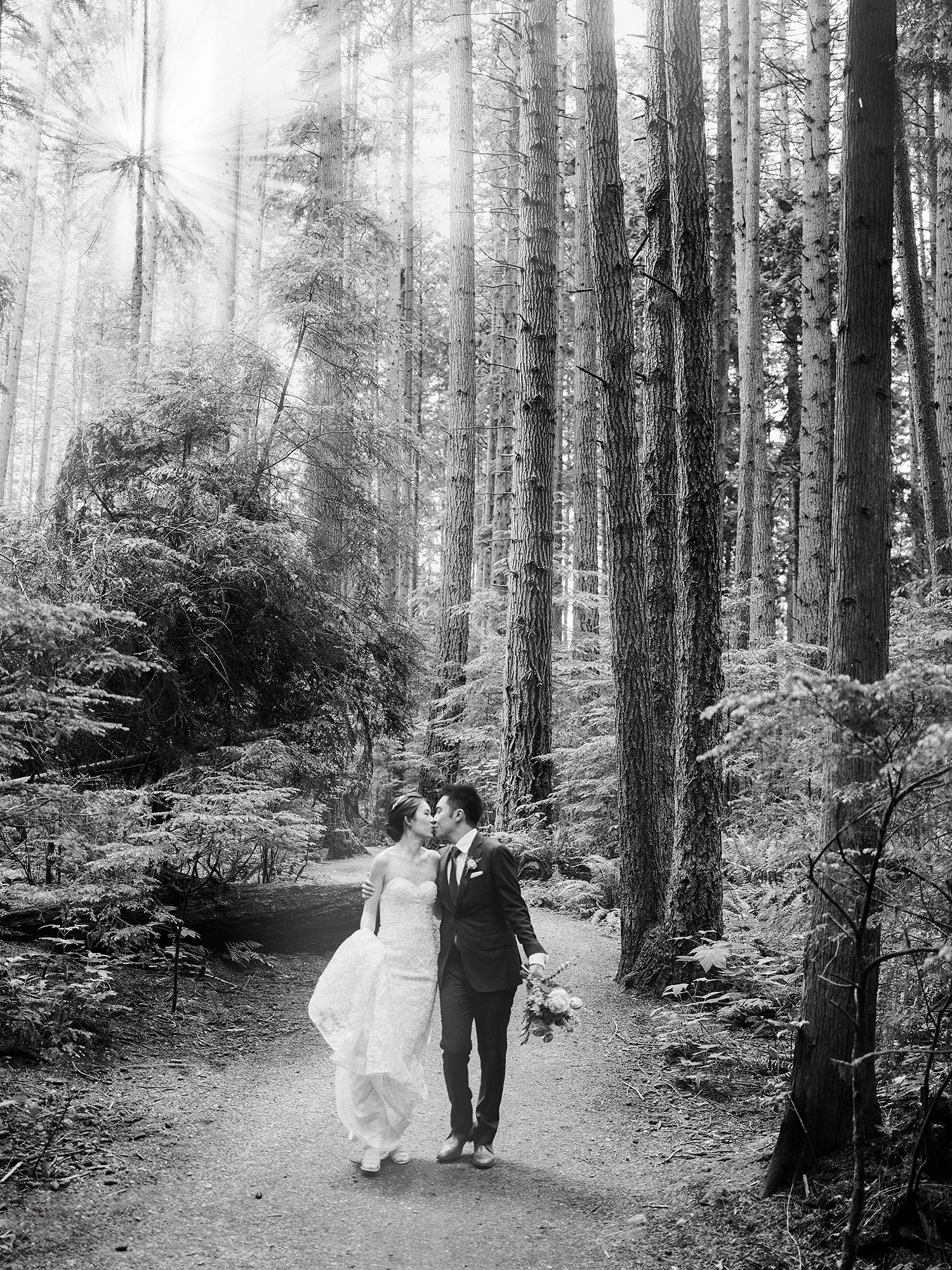 A couple in wedding attire walks through a misty forest path surrounded by tall evergreen trees. NYC wedding photographer.