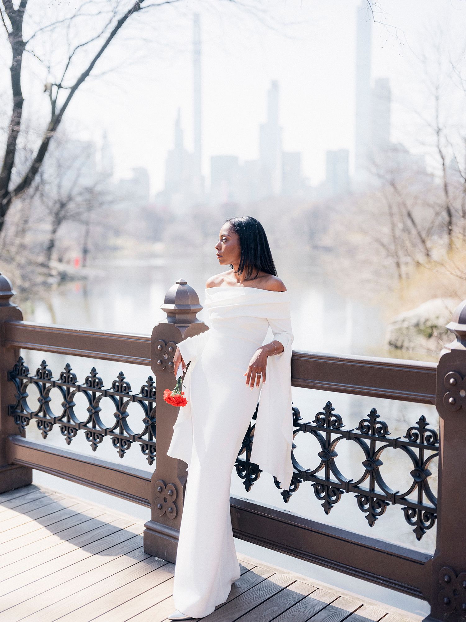 Elegant bride in white off-shoulder dress poses on bridge with NYC skyline in background. Central park, NYC wedding photographer.