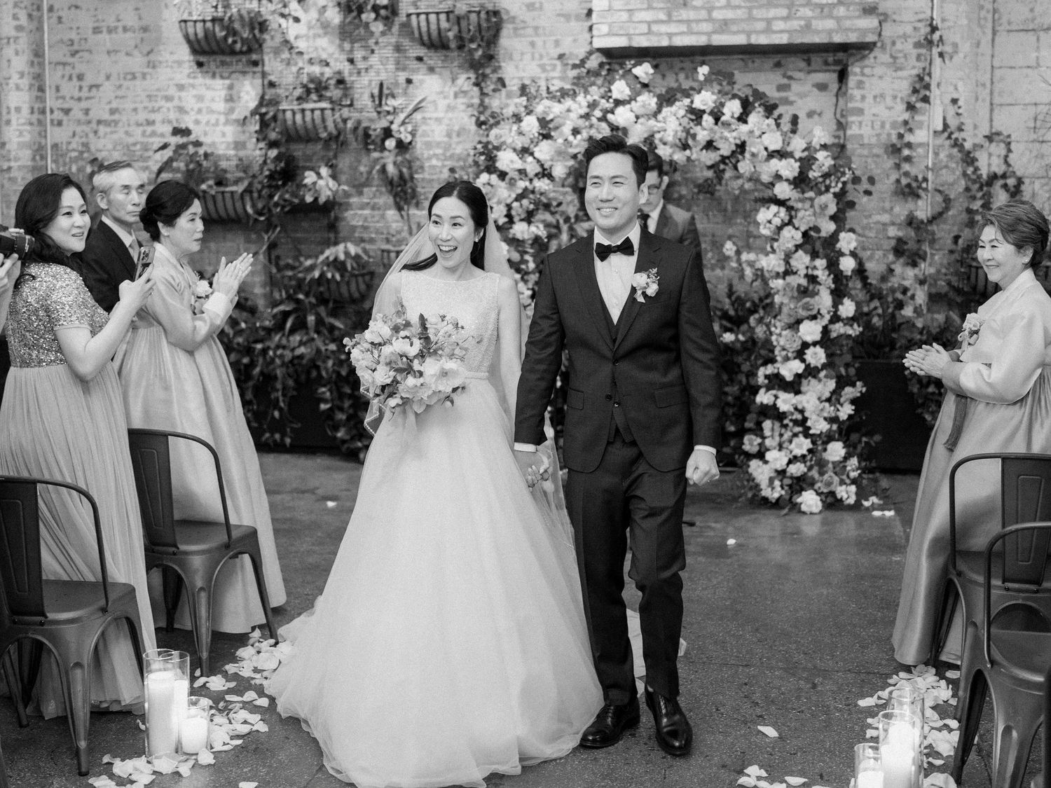 Wedding party gathered in front of a rustic brick wall decorated with elegant floral arrangements.