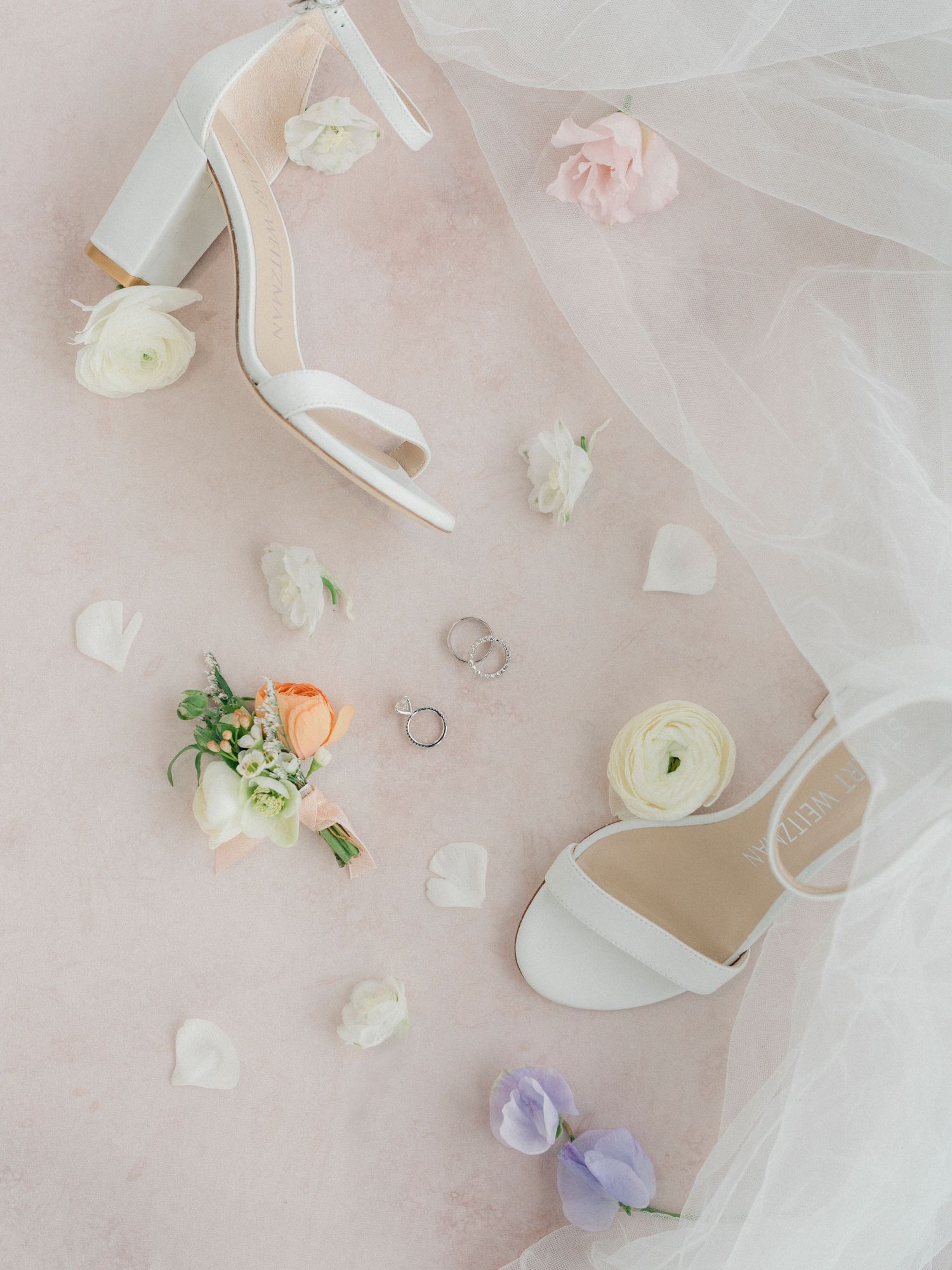 Elegant white bridal shoes with wedding rings and delicate flower petals on pink background.