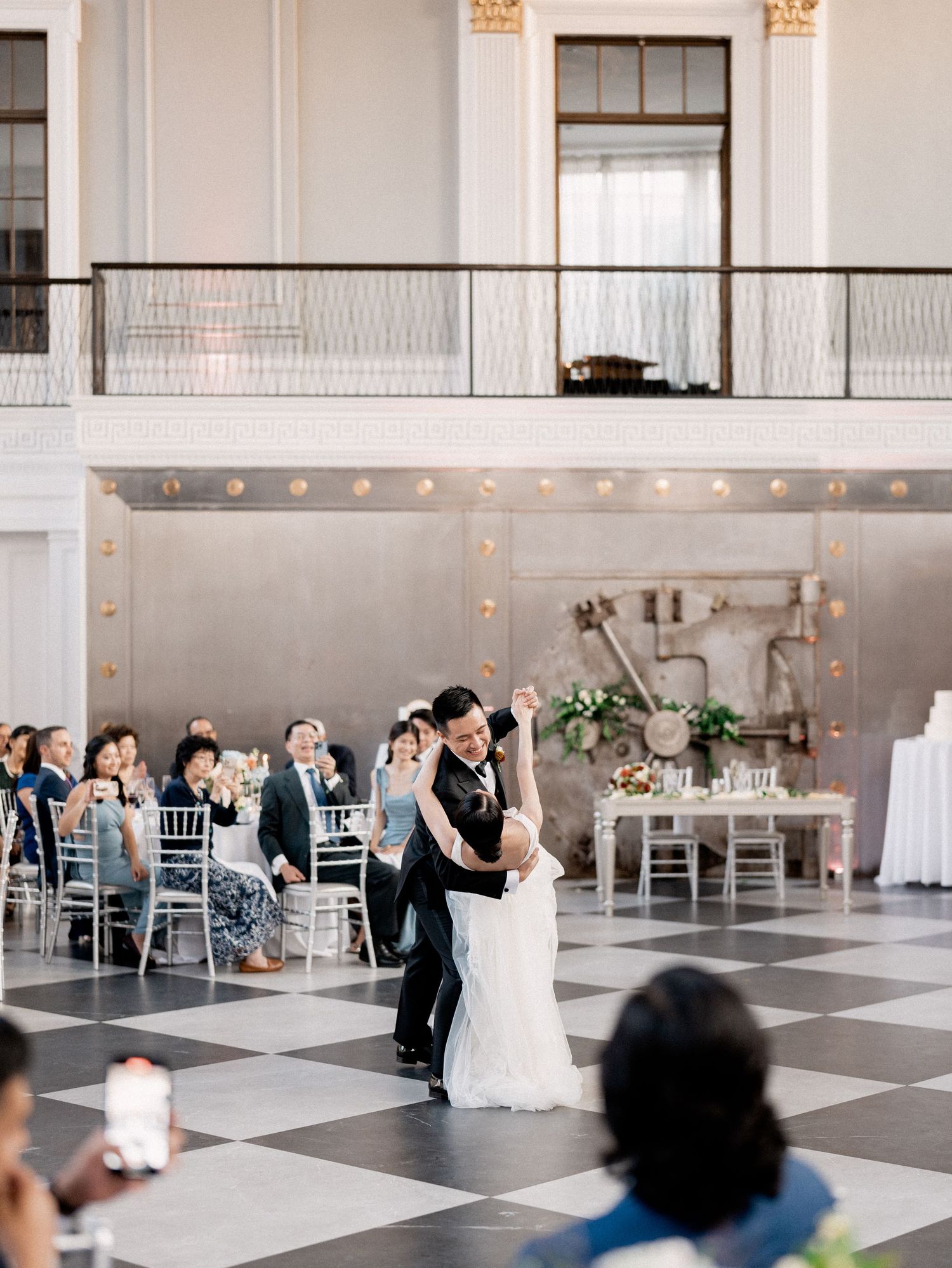 Couple dancing together in an elegant white reception venue with checkerboard floor and silver chiavari chairs.