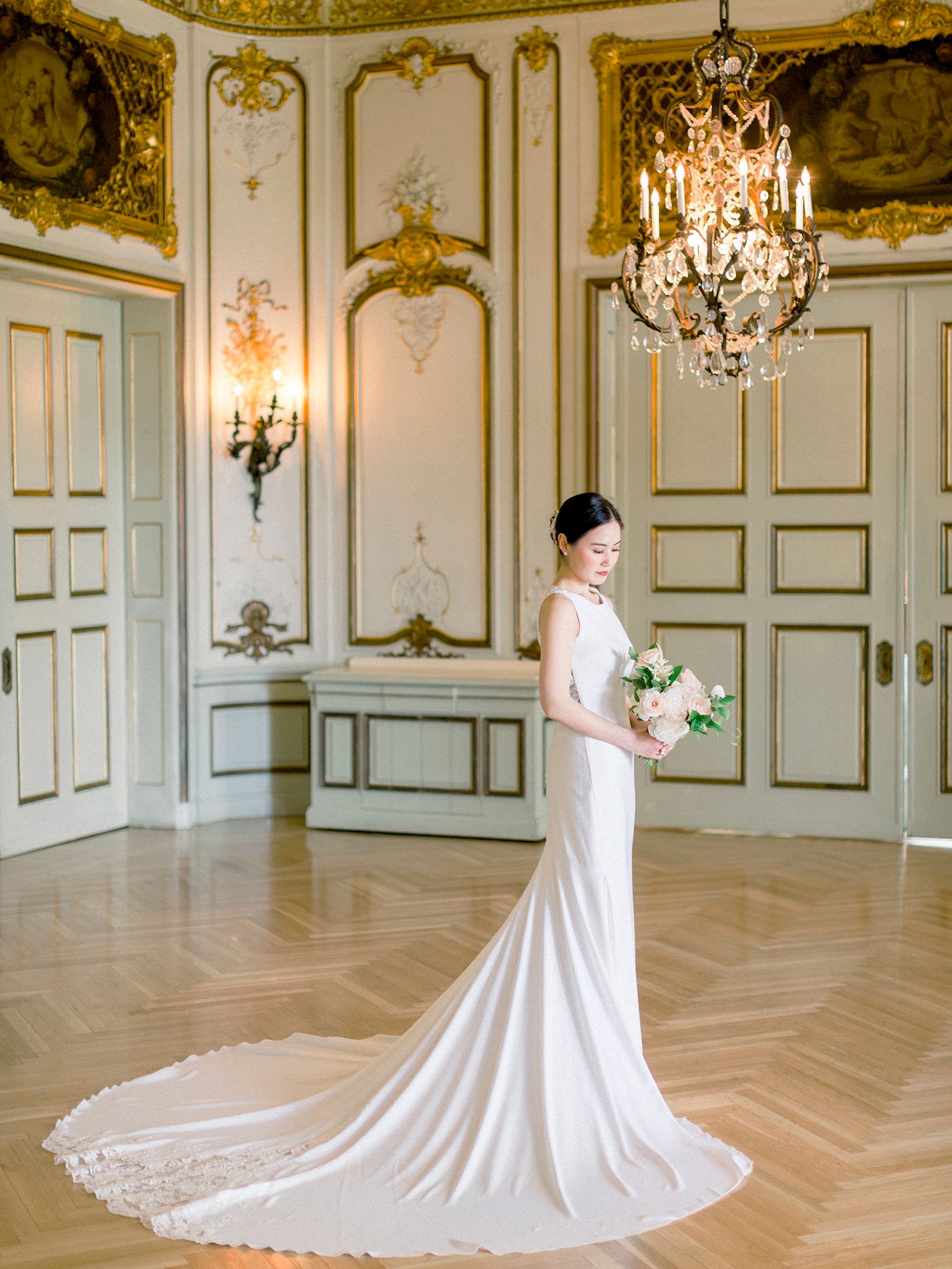 Bride in flowing white gown photographed in ornate baroque-style room with crystal chandeliers.
