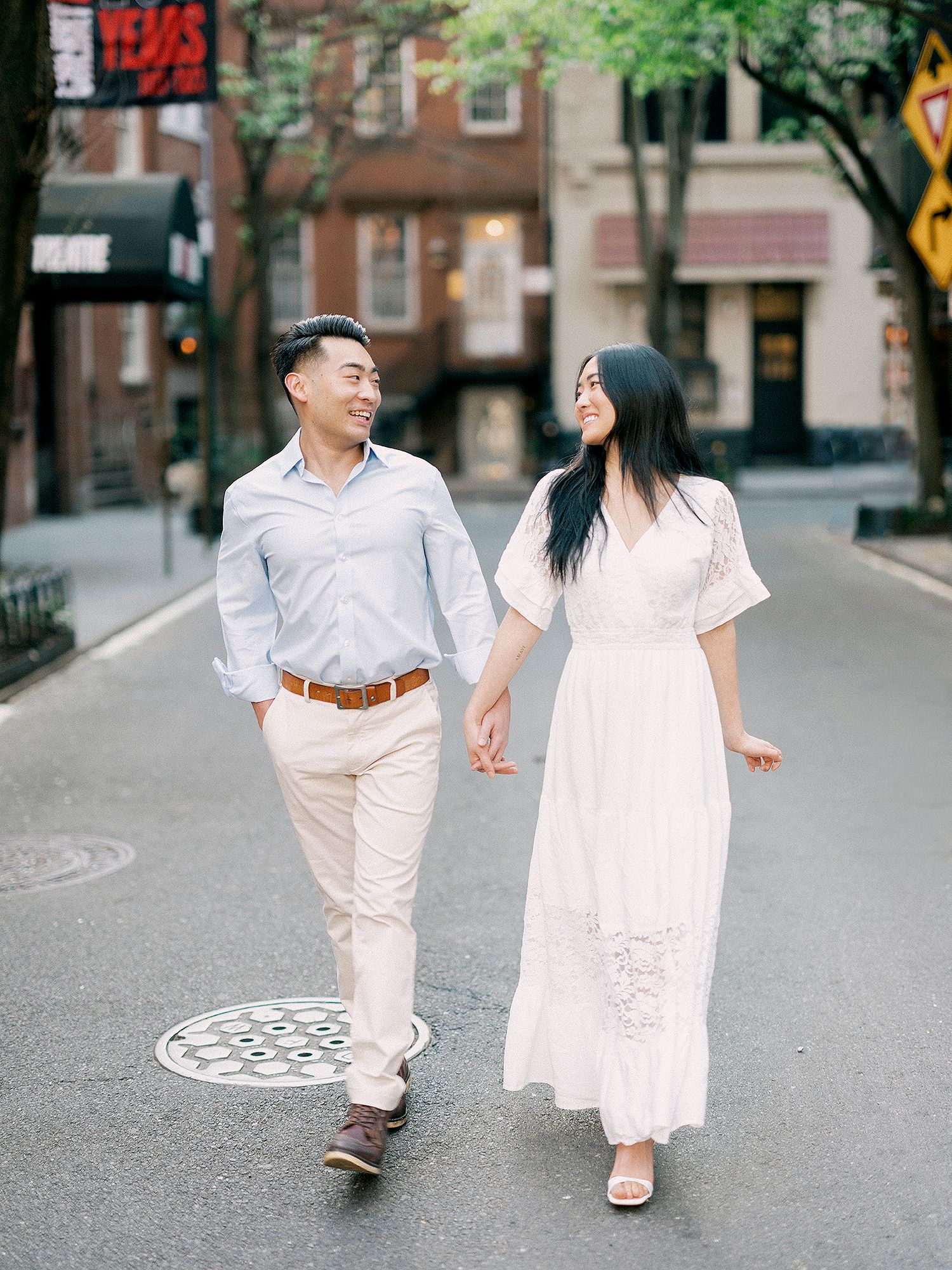 A couple in white summer outfits walks together on a quiet city street.
