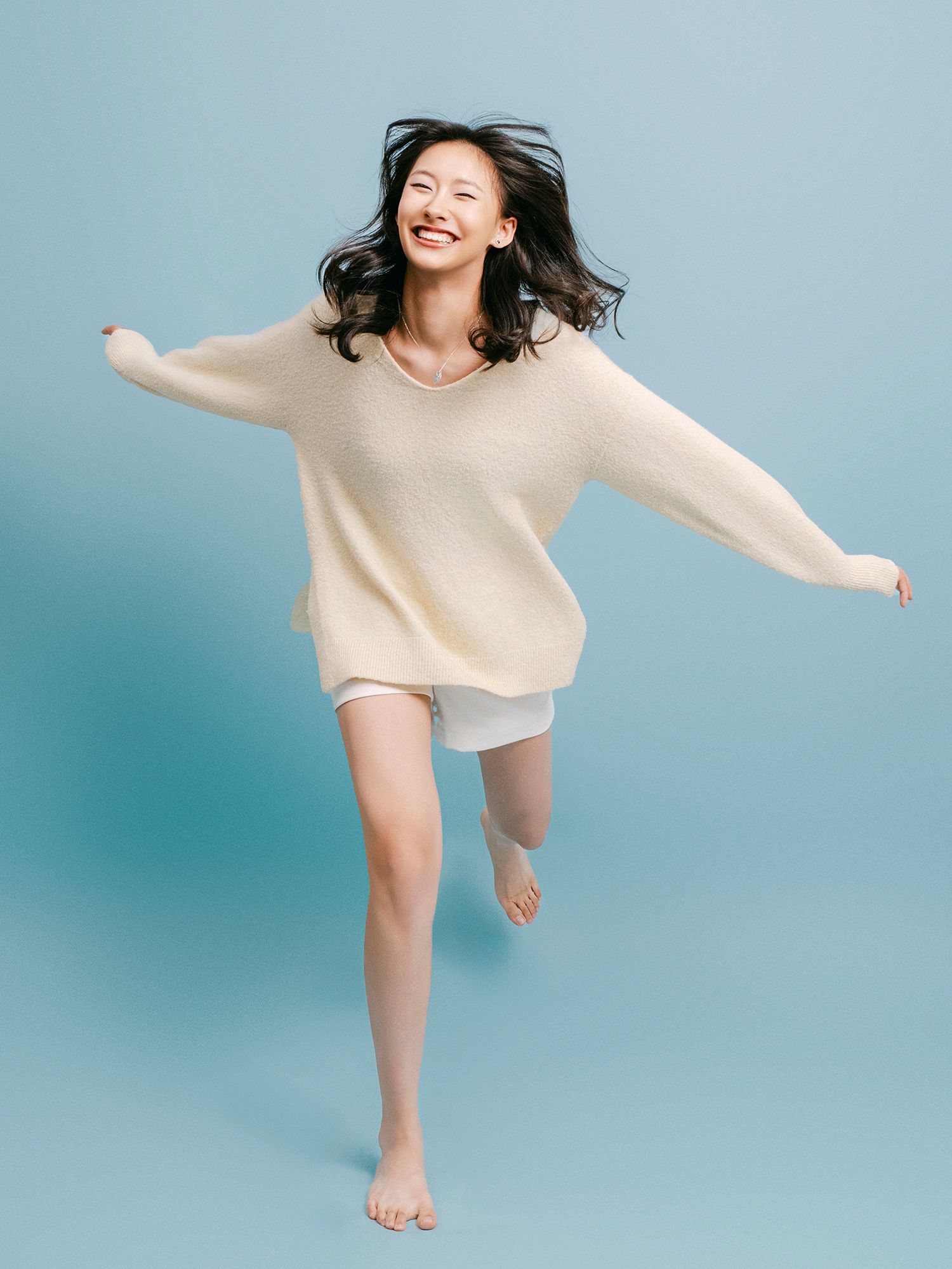 A person in a beige sweater jumps and dances joyfully against a light blue studio backdrop.