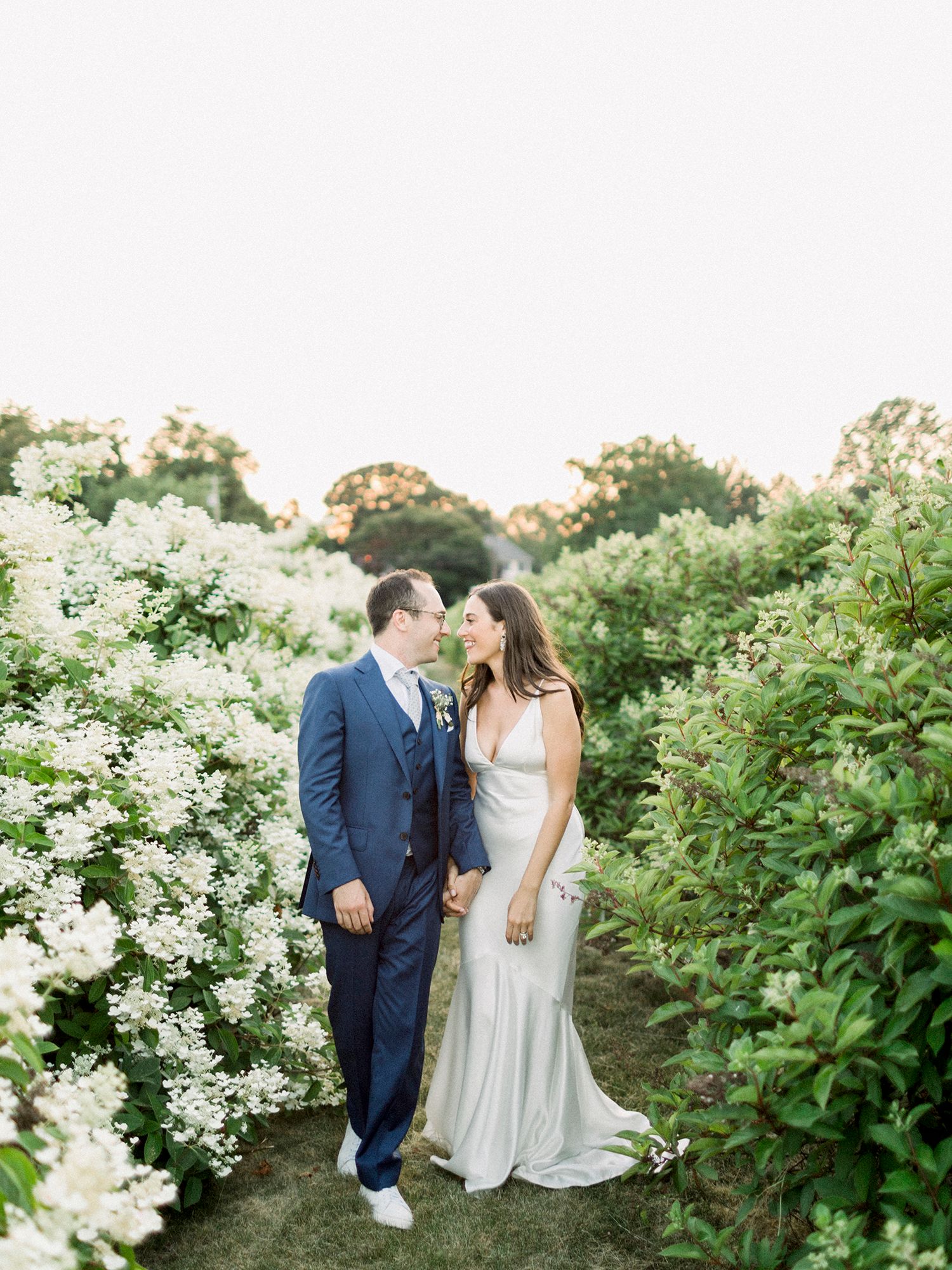 A couple walks through a beautiful garden path lined with white flowering bushes.