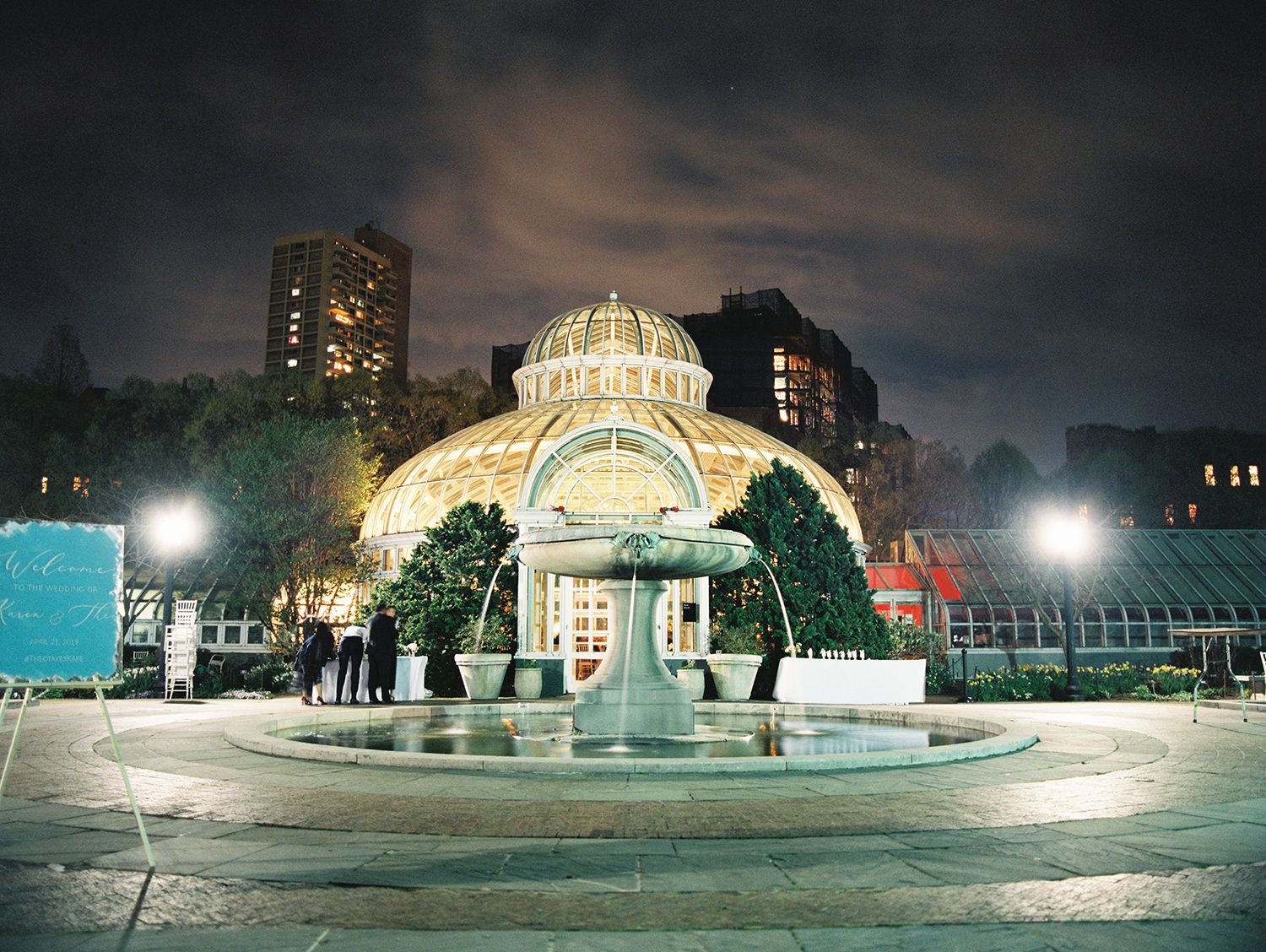 A panoramic nighttime view of an illuminated fountain and domed structure in a city park with glowing lights.