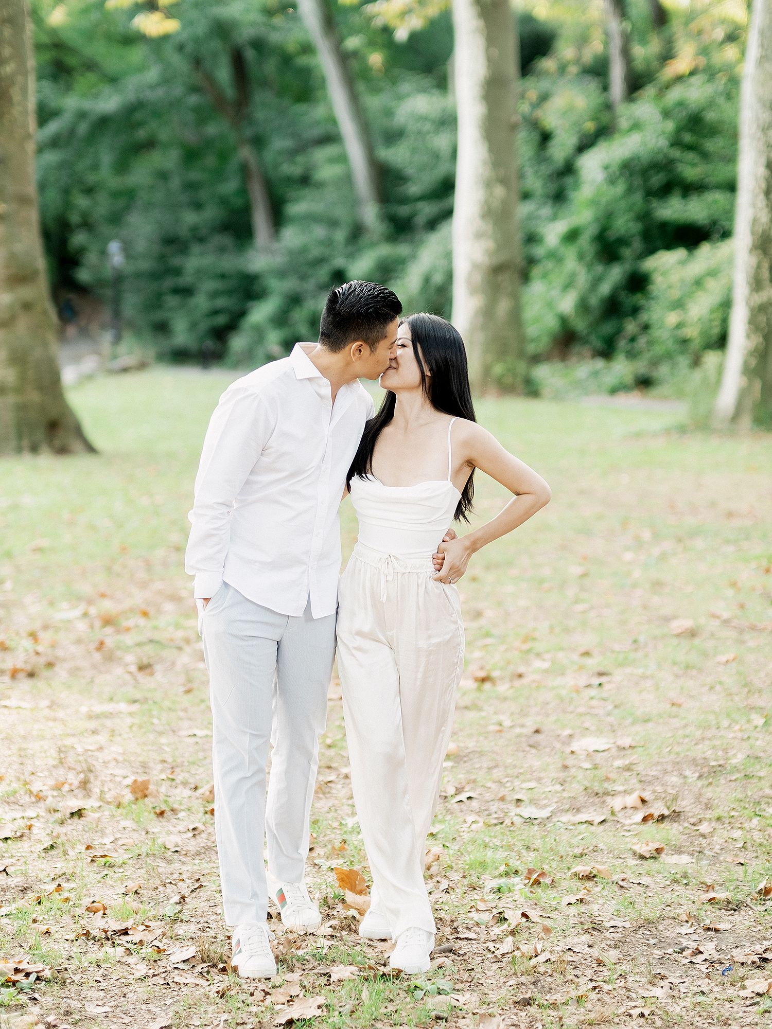 A couple in white outfits walks together through a sunlit wooded area.