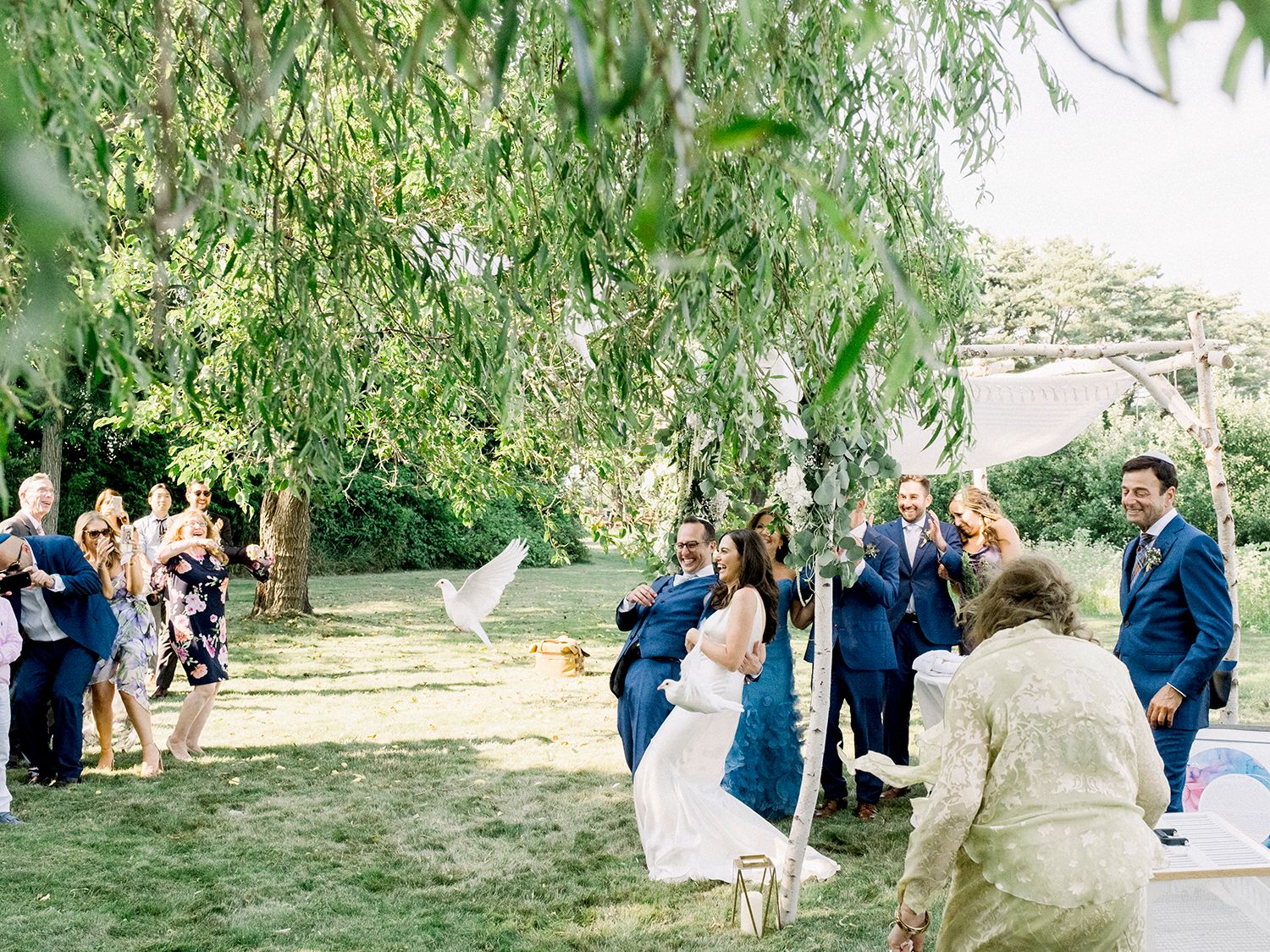 Wedding guests celebrate during outdoor ceremony under weeping willow trees.