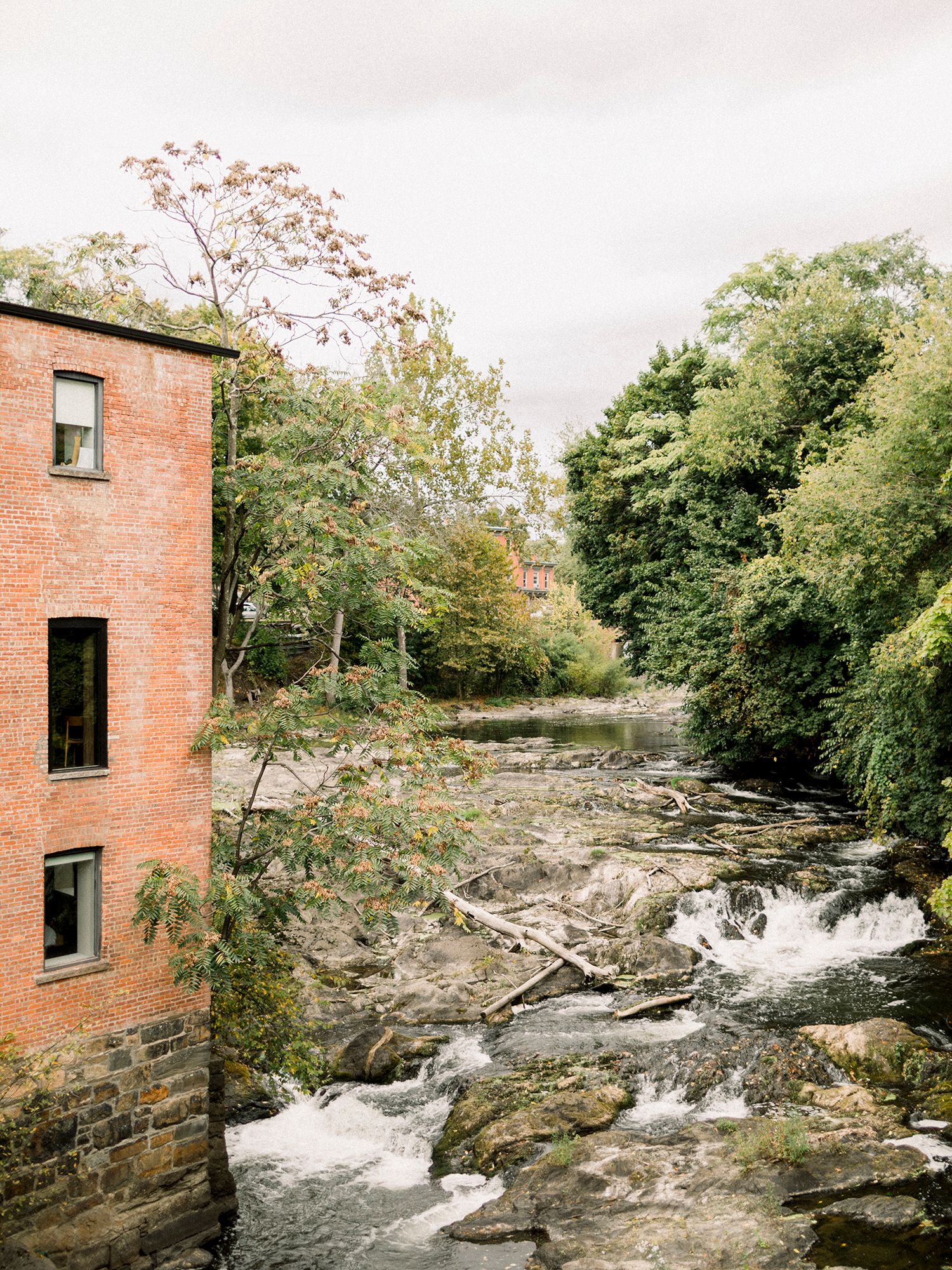Old brick mill building beside rushing waterfall and rocky riverbed.