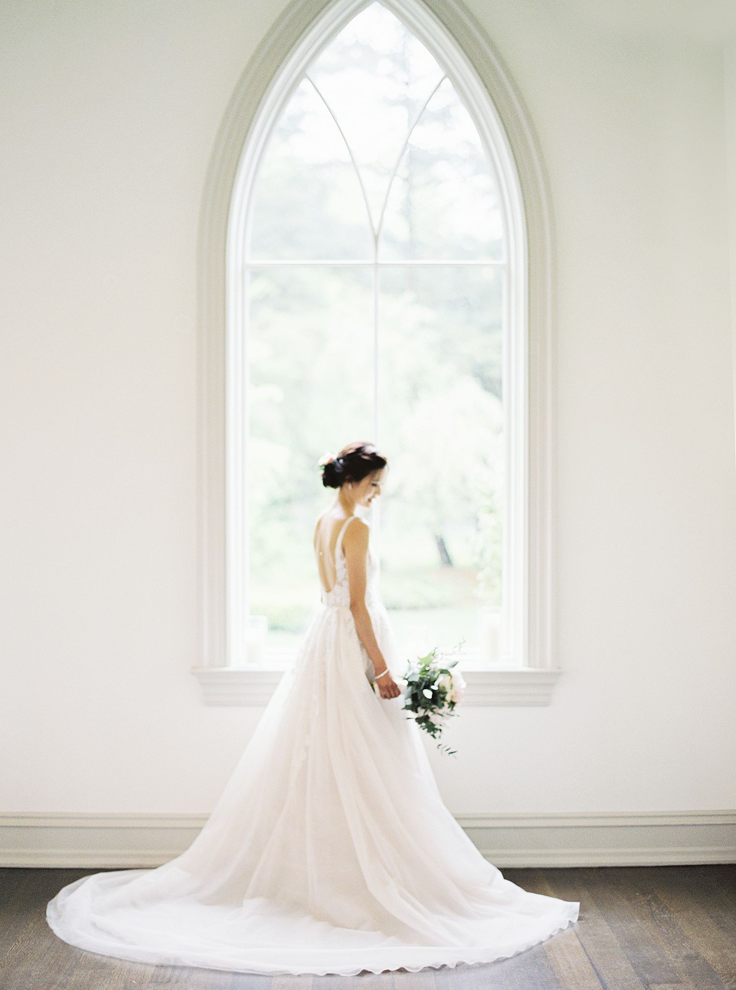 A white wedding gown with a flowing train is photographed in front of an arched church window. NYC wedding photographer.
