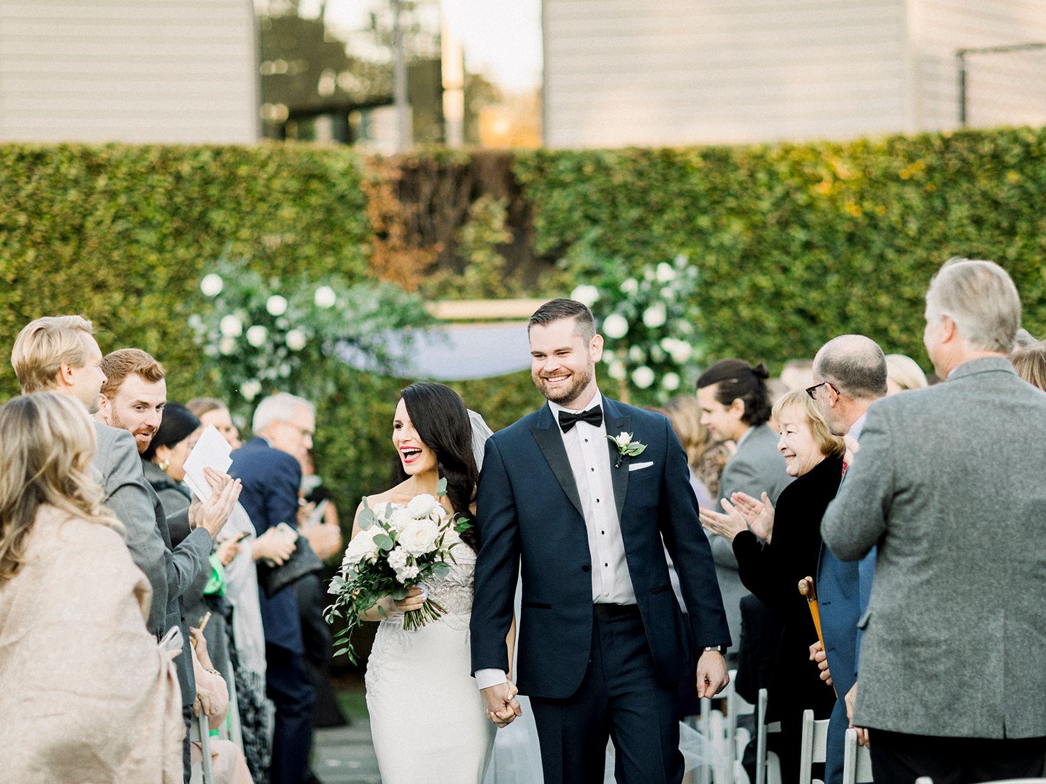 Wedding celebration on a lawn with guests mingling near a white building and hedge wall.
