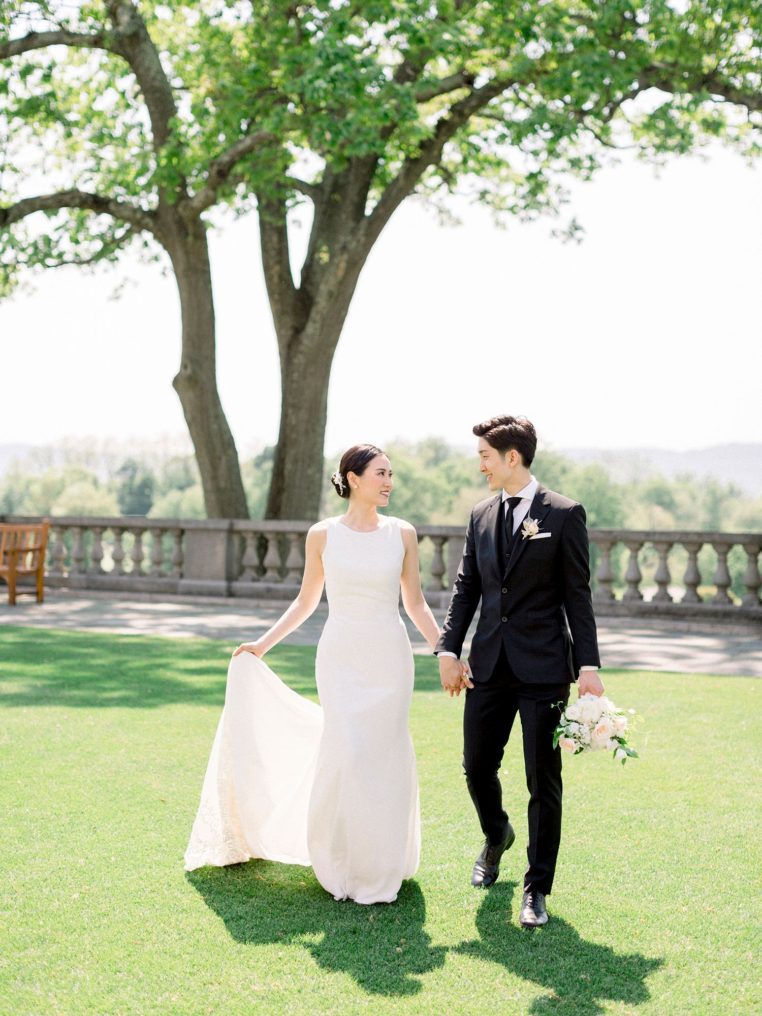 Couple strolls hand in hand on manicured lawn with grand tree and stone balustrade backdrop.