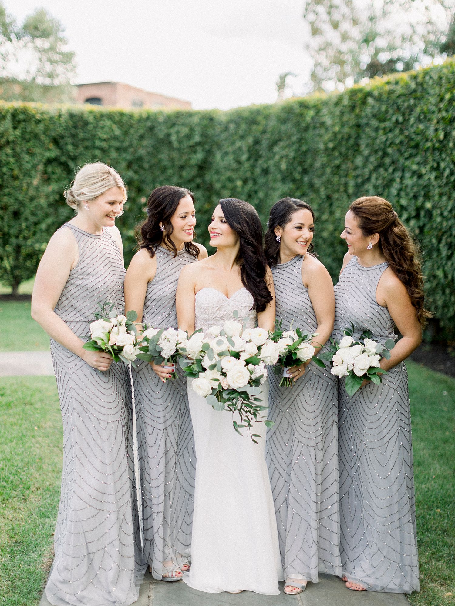 Wedding party in silver sequined dresses poses with white bouquets against a manicured hedge backdrop.
