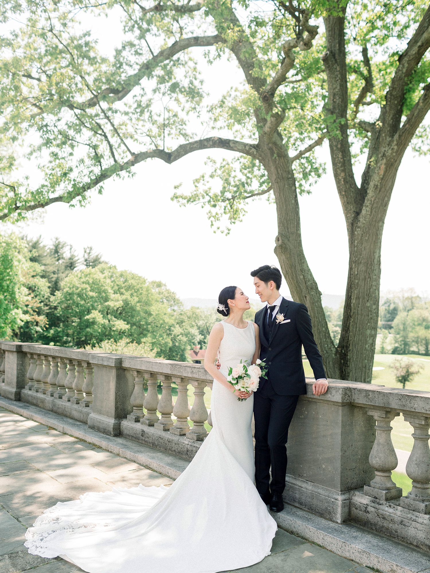 Romantic wedding portrait on stone balustrade with blooming trees creating natural canopy.