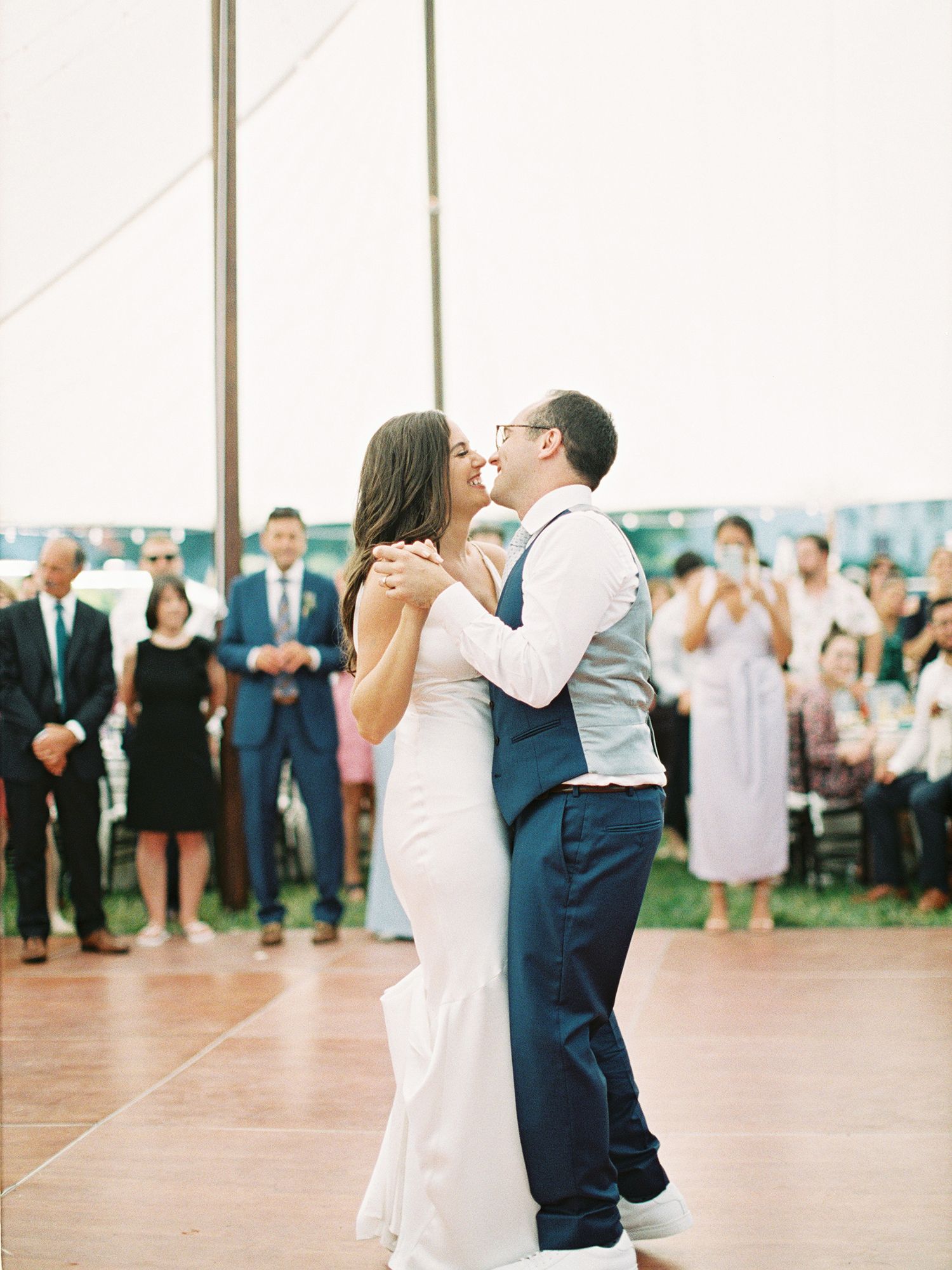 A couple shares their first dance at a wedding reception under a white tent surrounded by guests. NYC wedding photographer.