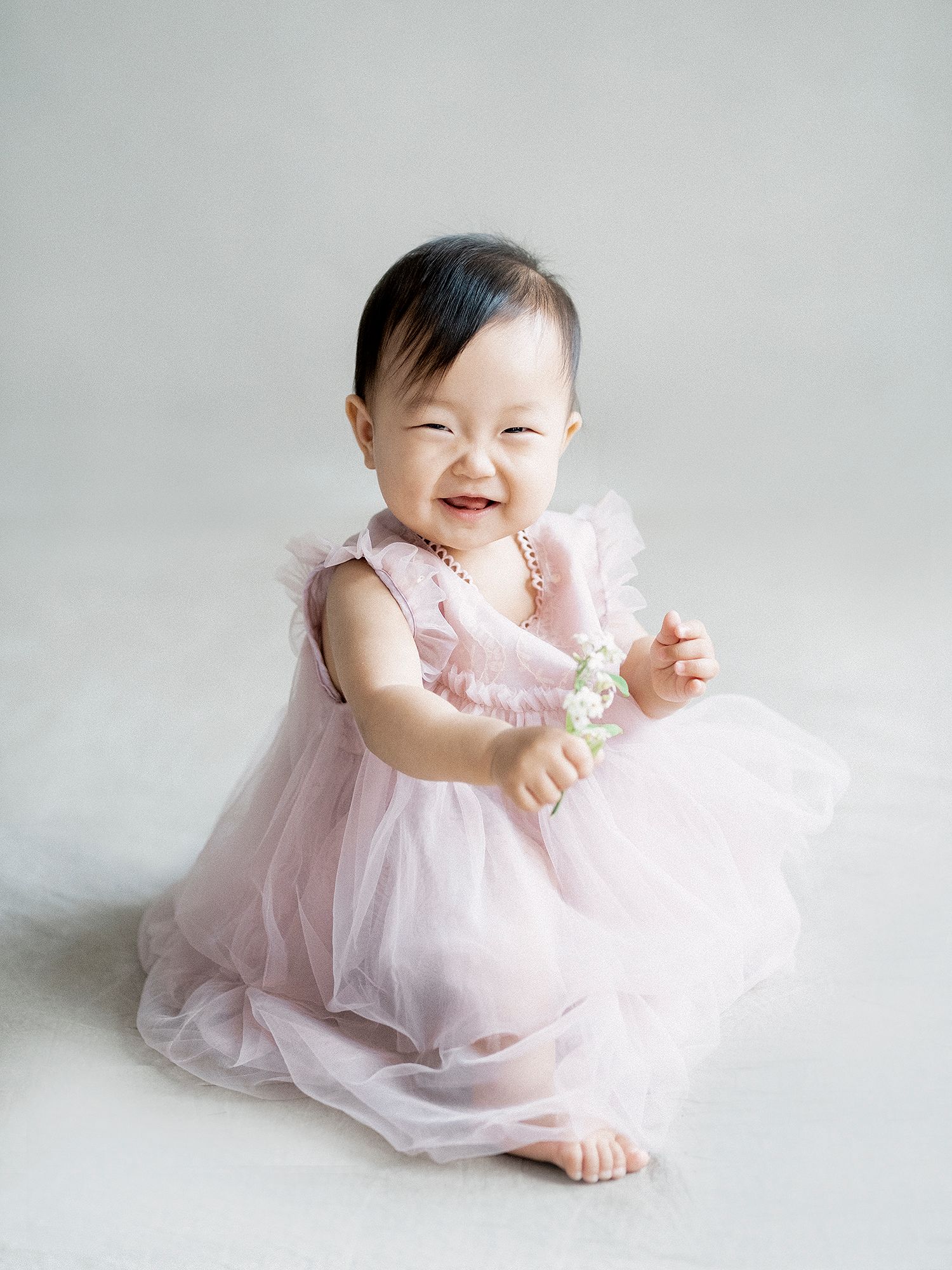 A baby in a pink tulle dress sits and smiles against a light gray background in a series of sweet poses.