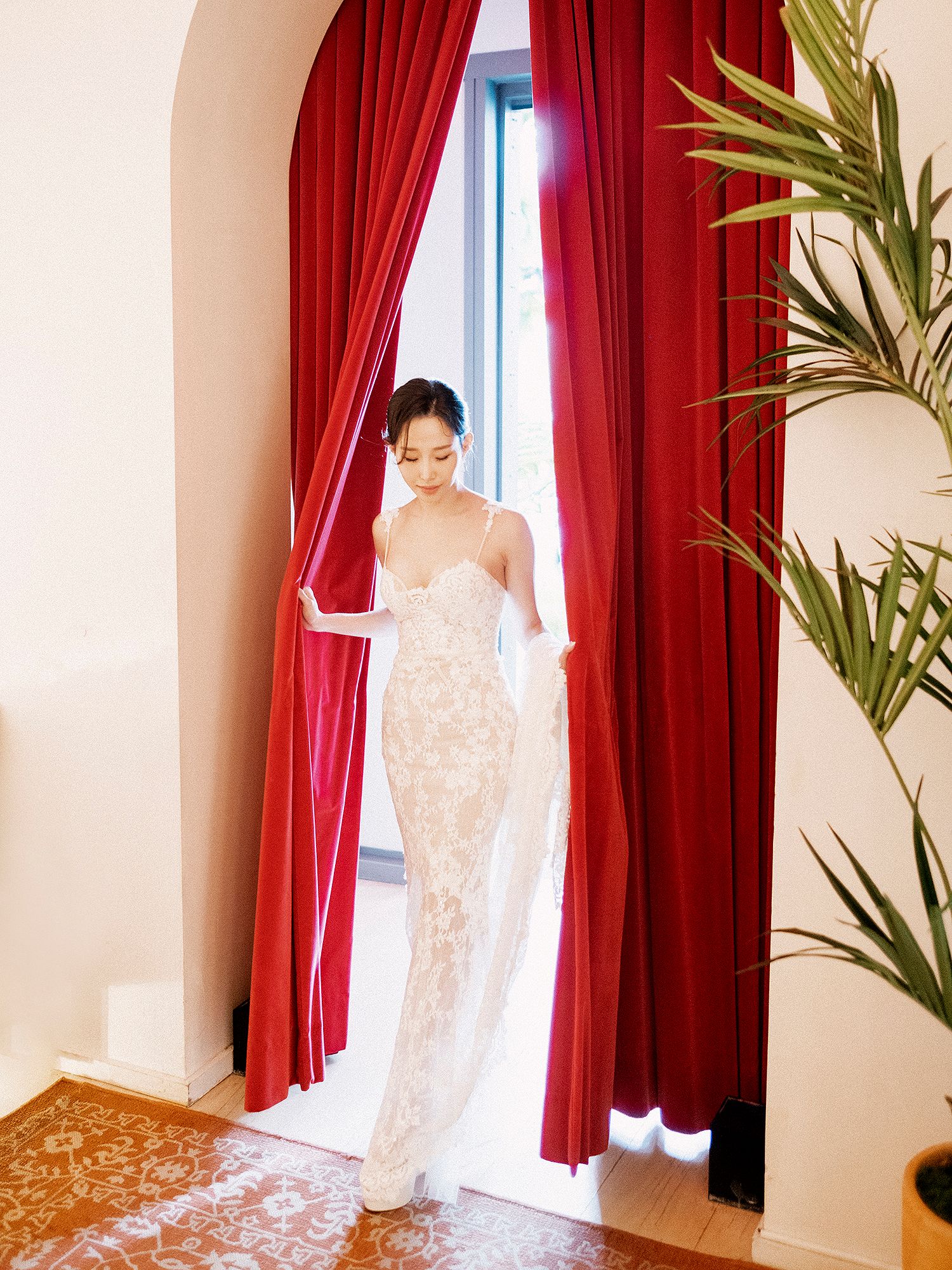 A wedding dress is displayed against red curtains and tropical plants in an elegant room setting. NYC wedding photographer.