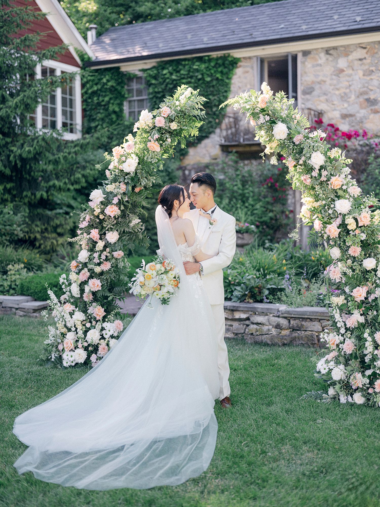 Couple shares intimate moment under floral arch with blush and white roses against stone house. NYC wedding photographer.