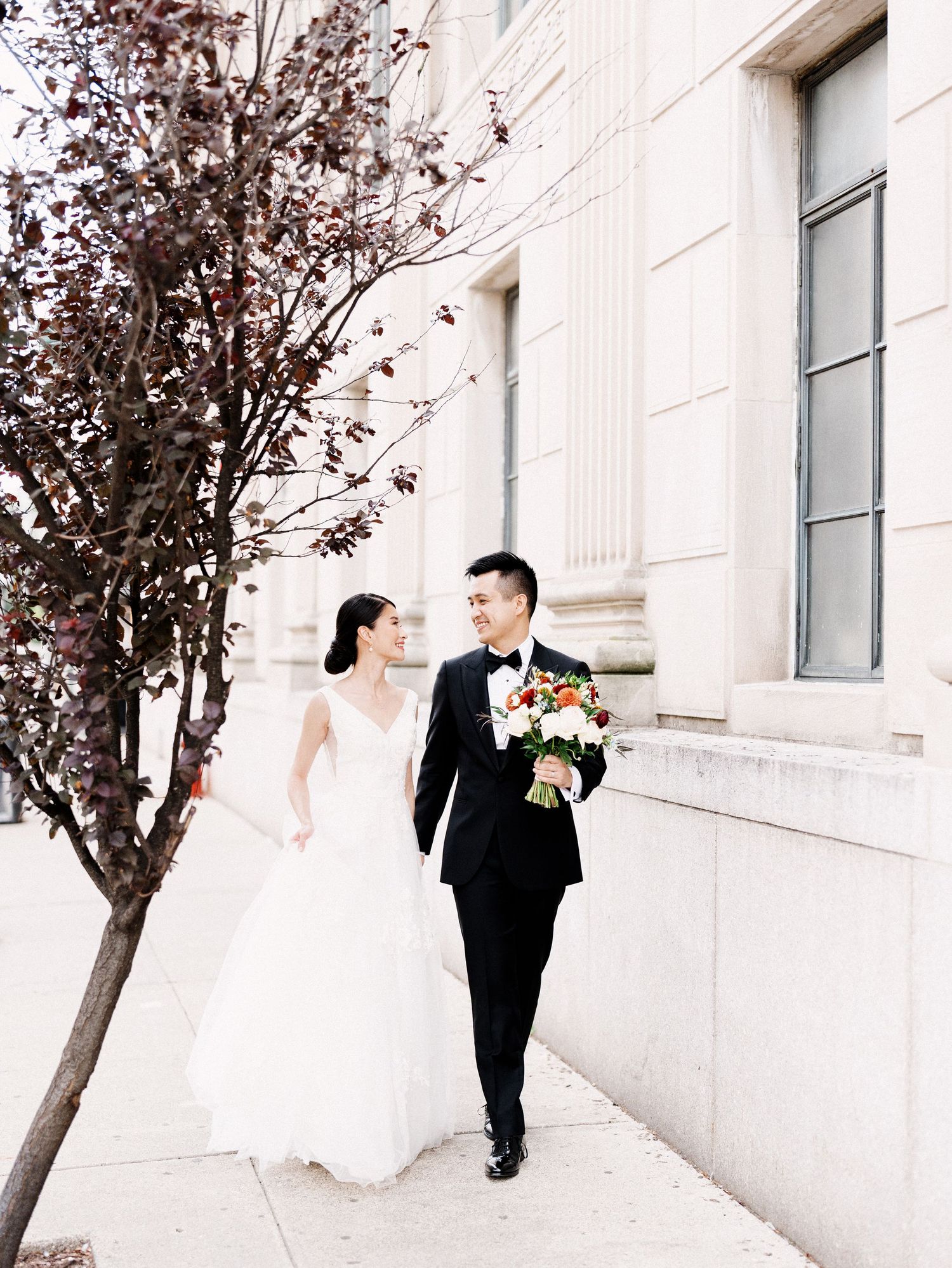 Elegant wedding couple walking along classic white stone building exterior.