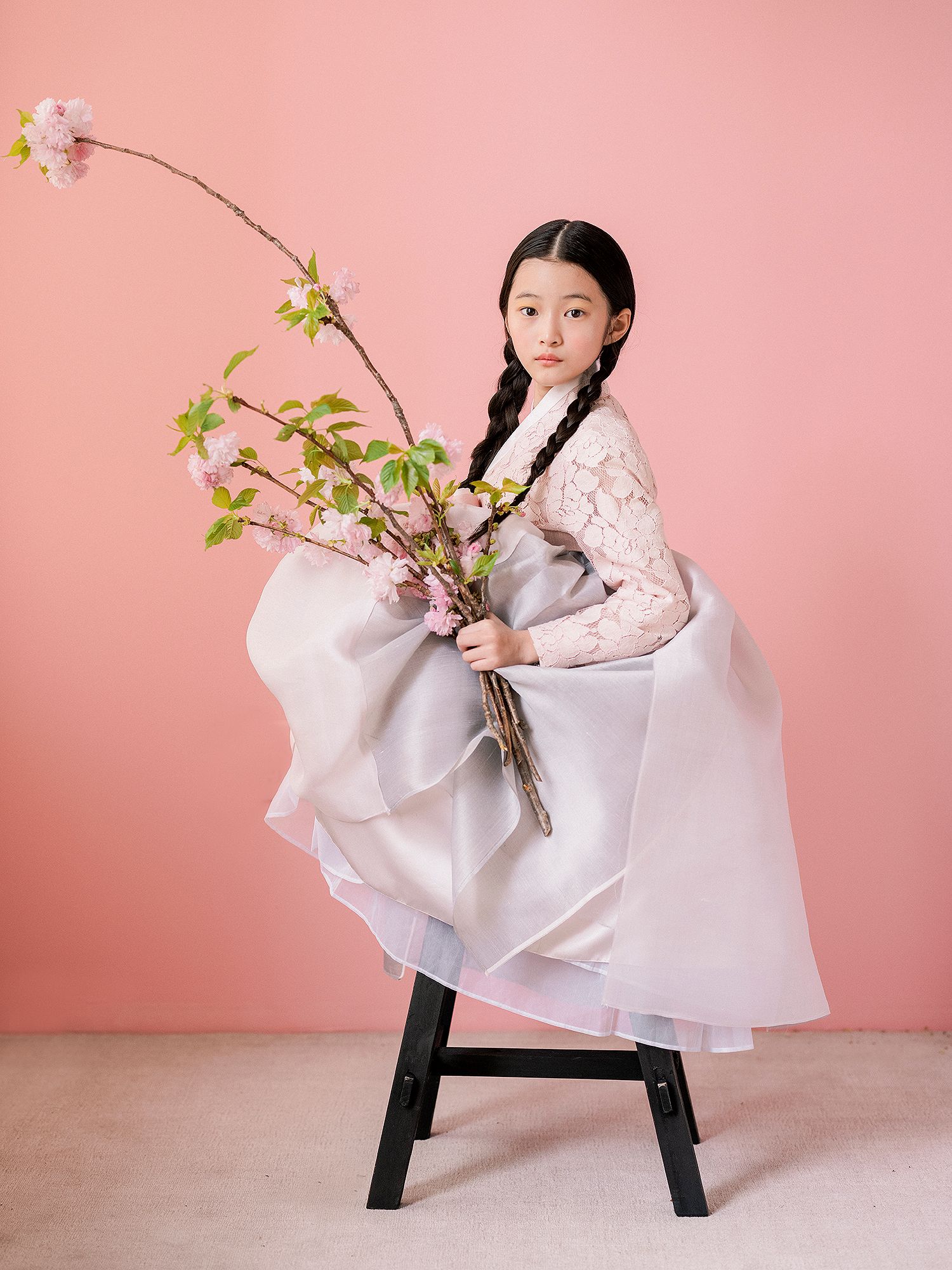 A child in a flowing white dress poses gracefully with cherry blossoms against a pink backdrop.