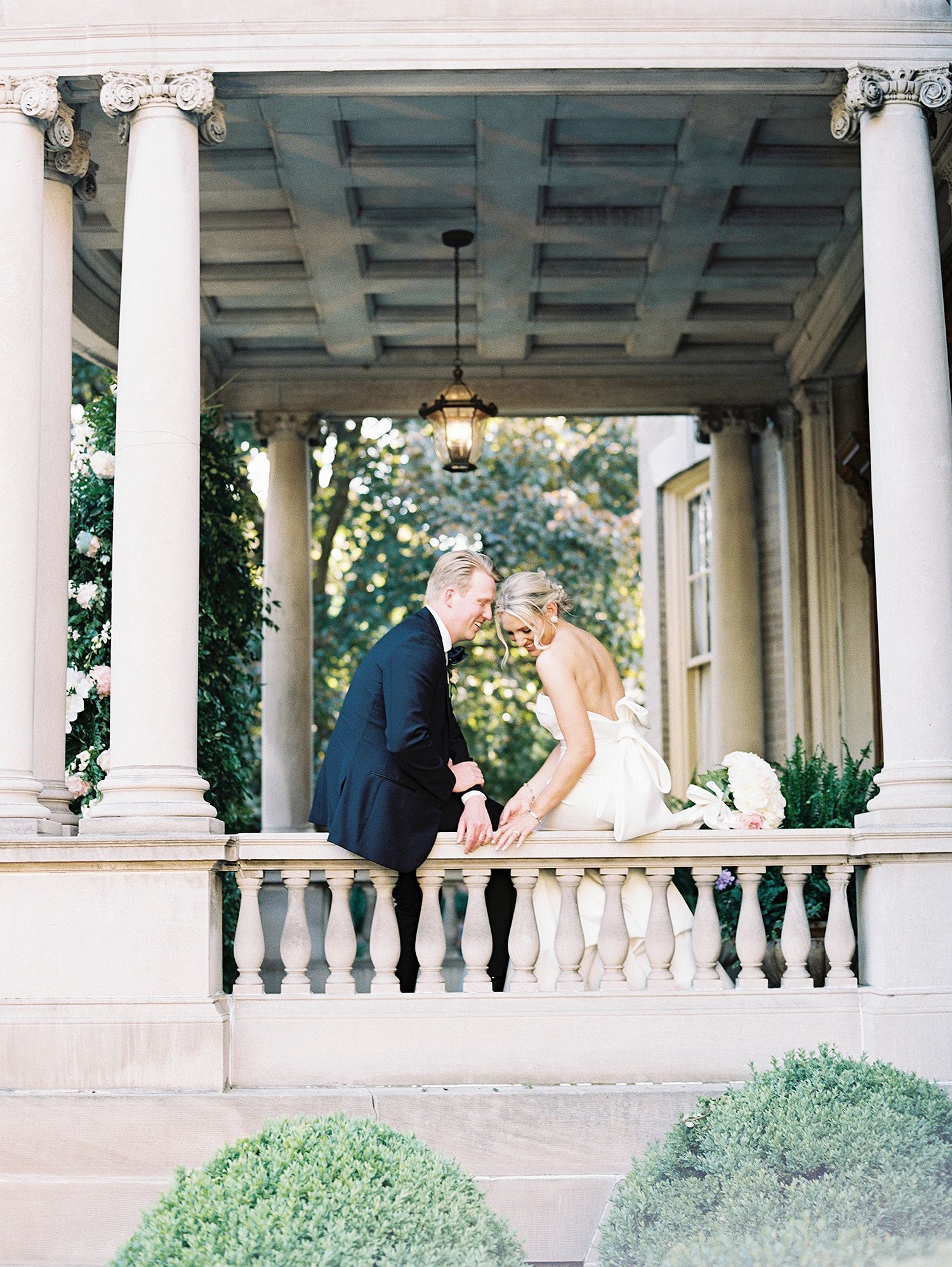 A wedding couple shares a romantic moment on a columned veranda with lush greenery in the background. NYC wedding photographer.