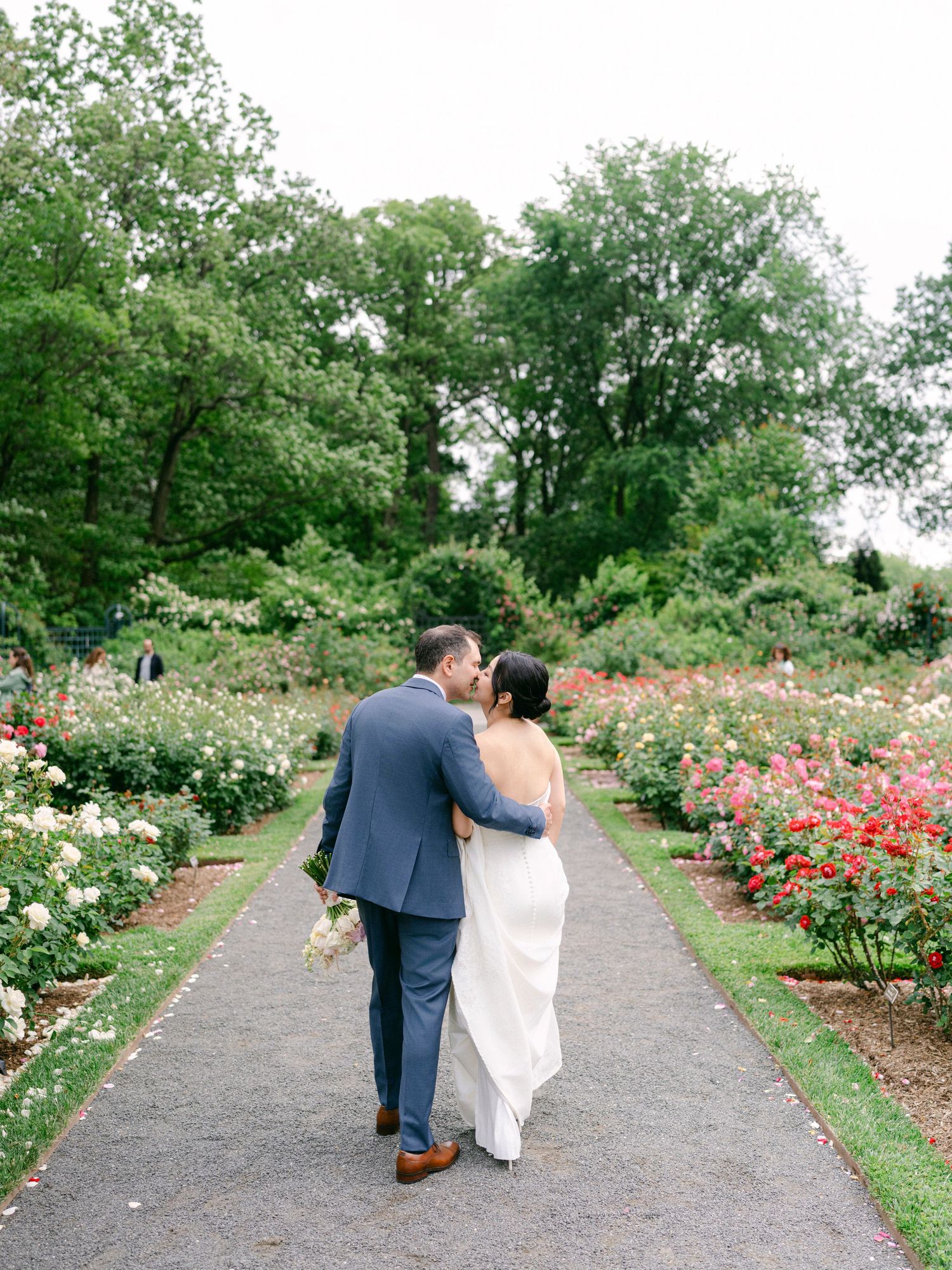 A couple walks along a garden path lined with blooming roses and lush greenery.