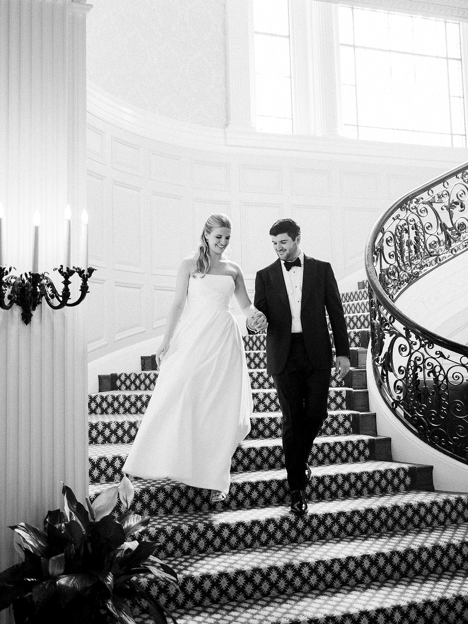 A couple descends an elegant grand staircase with ornate iron railings in formal attire at a luxurious venue. NYC wedding photographer.