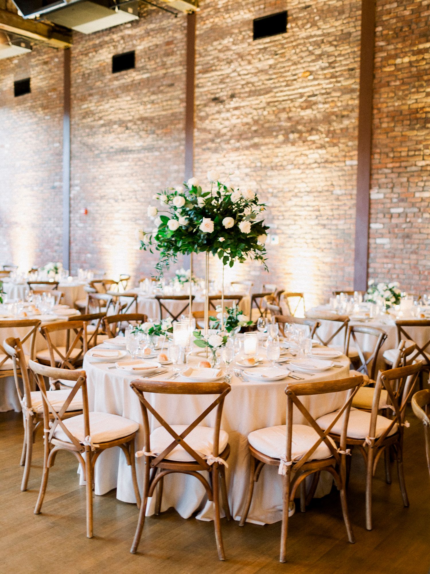 Wedding reception venue with exposed brick walls, white tablecloths, and rustic wooden chairs arranged for dining.
