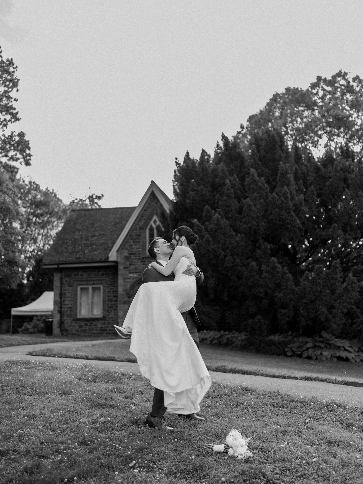 Black and white wedding photos showing couple dancing on grassy hill near rustic buildings.