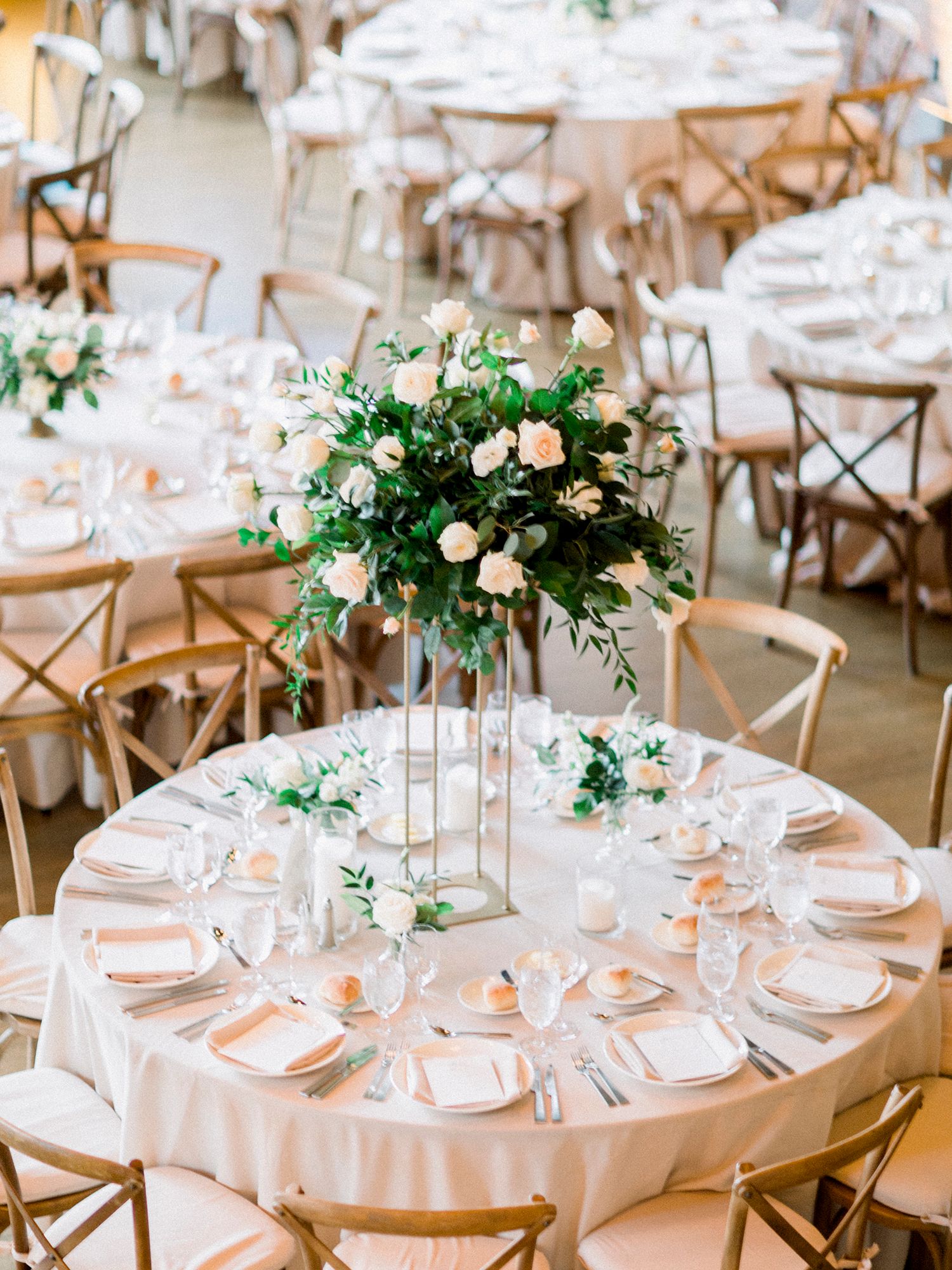 Elegant wedding reception tables set with white linens, floral centerpieces and wooden cross-back chairs in a bright venue.