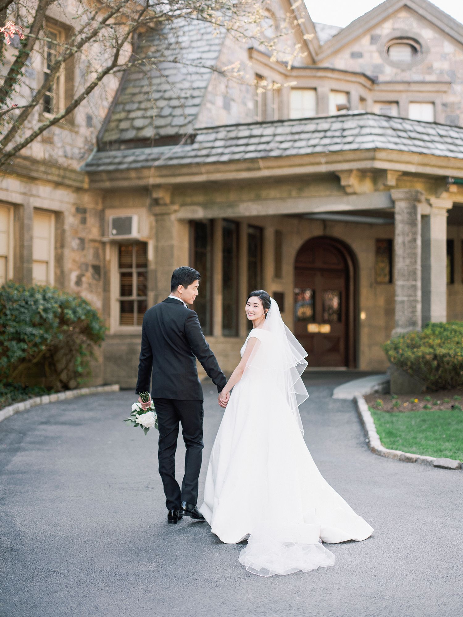A couple in wedding attire walks along a stone mansion's exterior path during their wedding day. NYC wedding photographer.