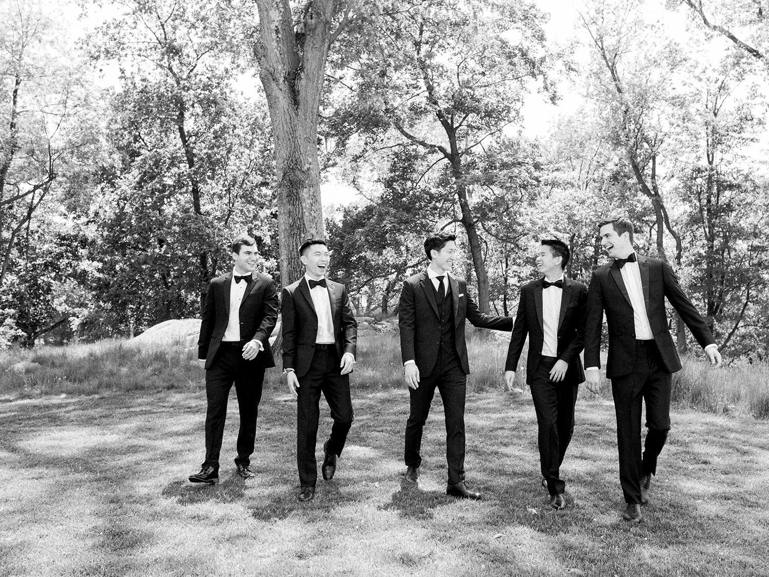 Wedding party poses in a casual line formation under mature trees in a black and white photograph.