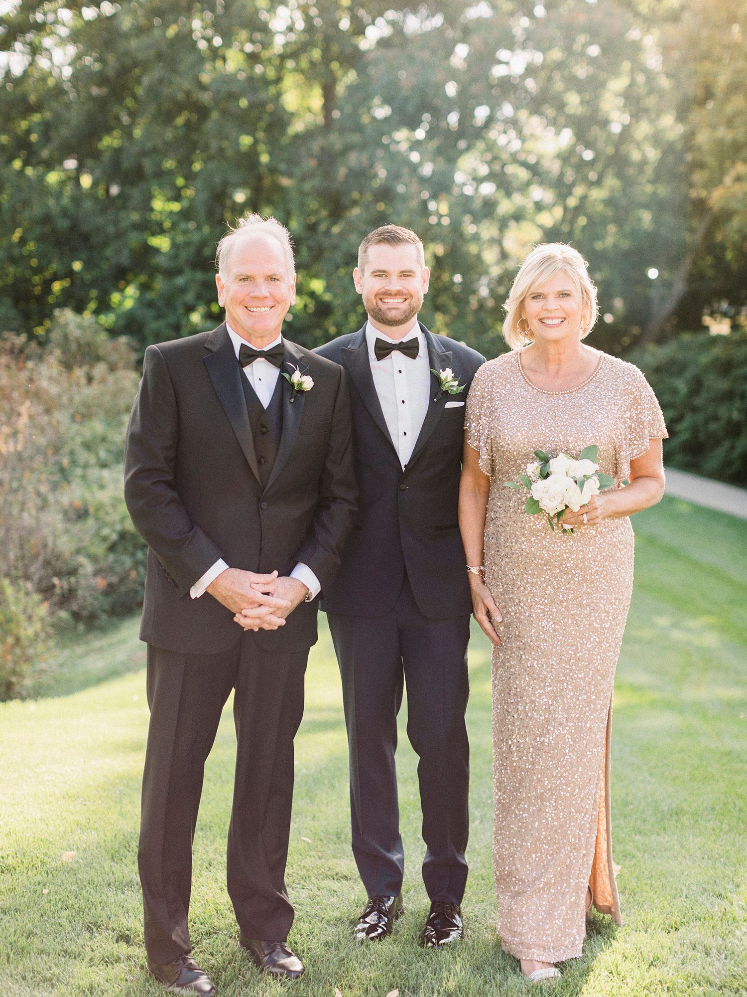 Wedding party members in formal attire pose together on a grassy lawn during golden hour.