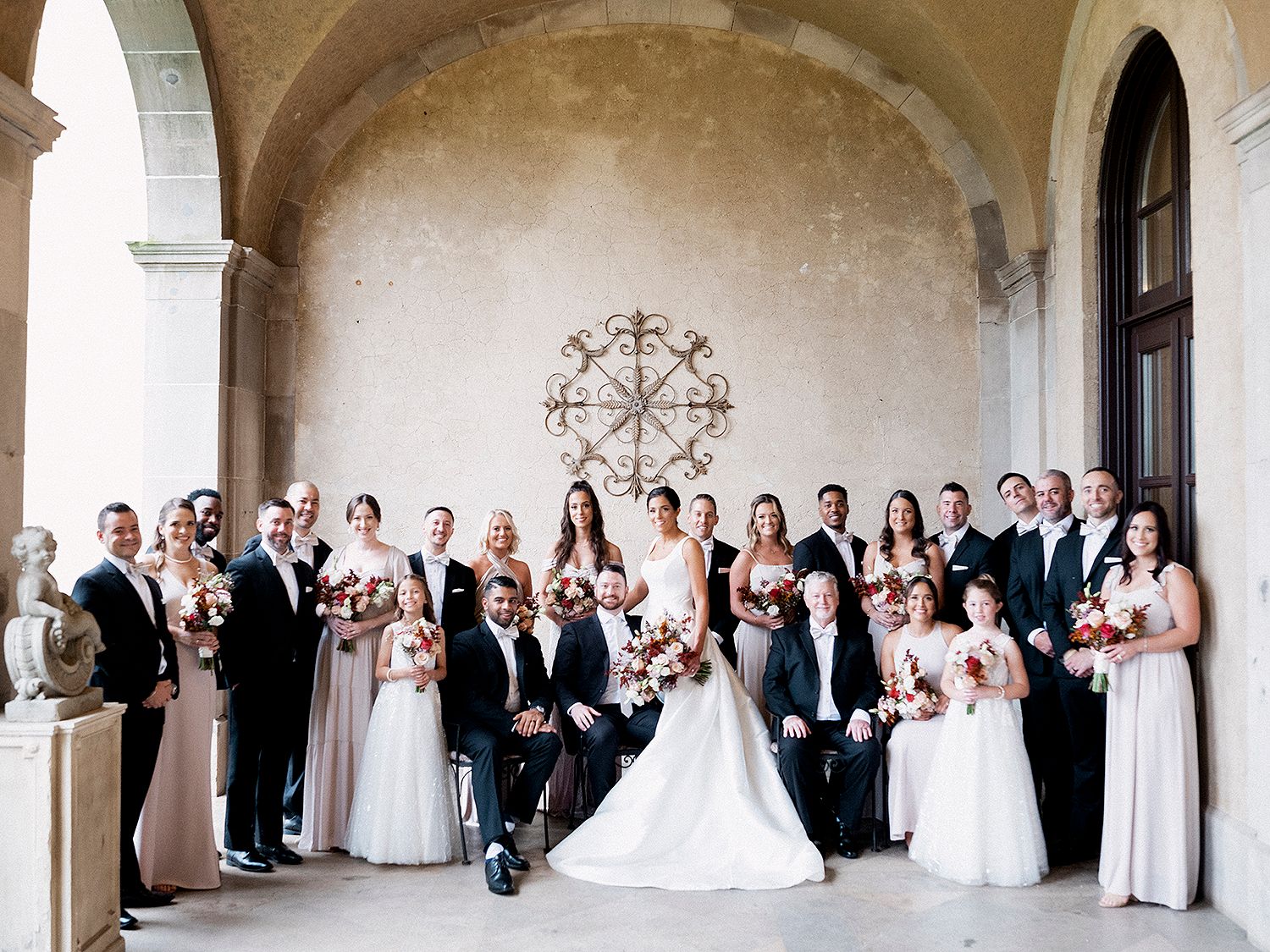 Large wedding party posed in formal attire under arched colonnade. NYC wedding photographer.