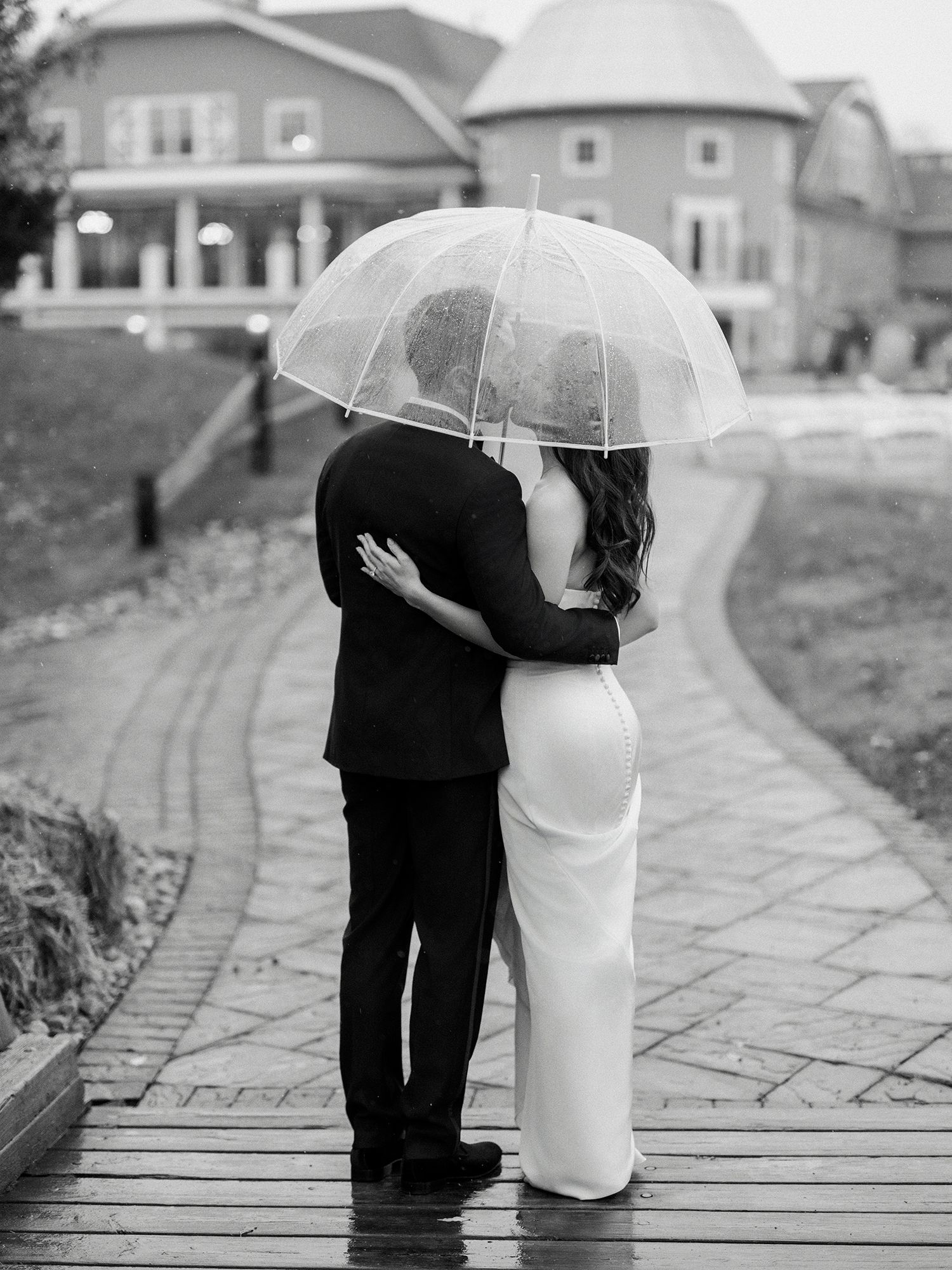 A couple shares an intimate moment under clear umbrellas on a stone pathway in black and white. NJ Wedding photographer.